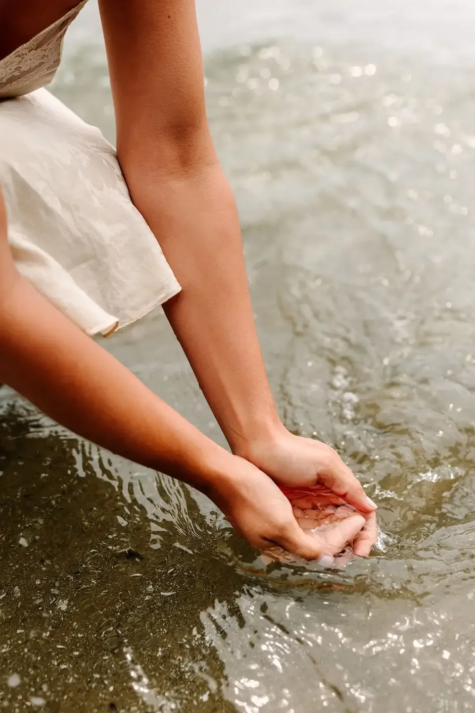 Hélène with tanned skin wearing a light-colored dress, crouching at the edge of the water on the beach, cupping their hands in the shallow ocean water.