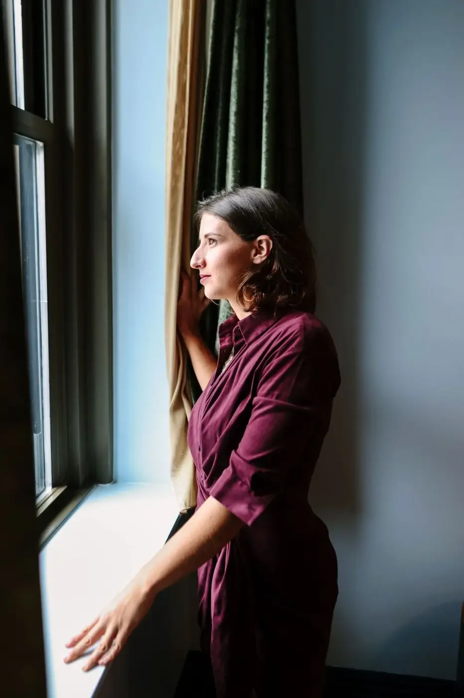 Hélène with shoulder-length dark hair standing by a window, looking outside, with her hand on the curtains, wearing a burgundy blouse.