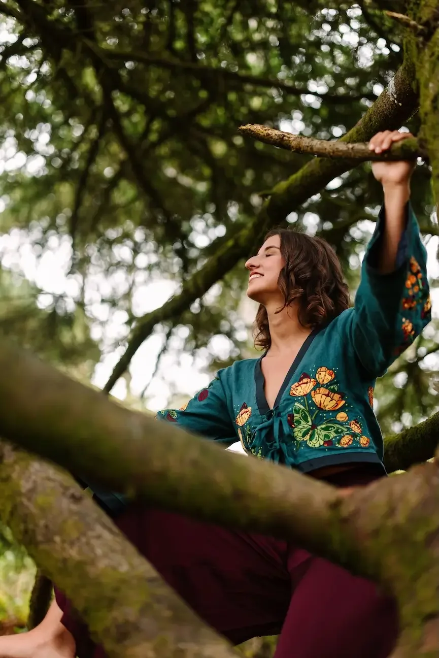 Hélène with brown, curly hair sitting cross-legged on a tree branch, smiling with her eyes closed, surrounded by green foliage.