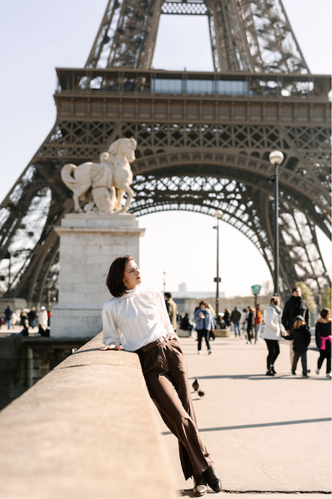 Hélène leaning against a stone railing near the Eiffel Tower in Paris, France, with people walking in the background.