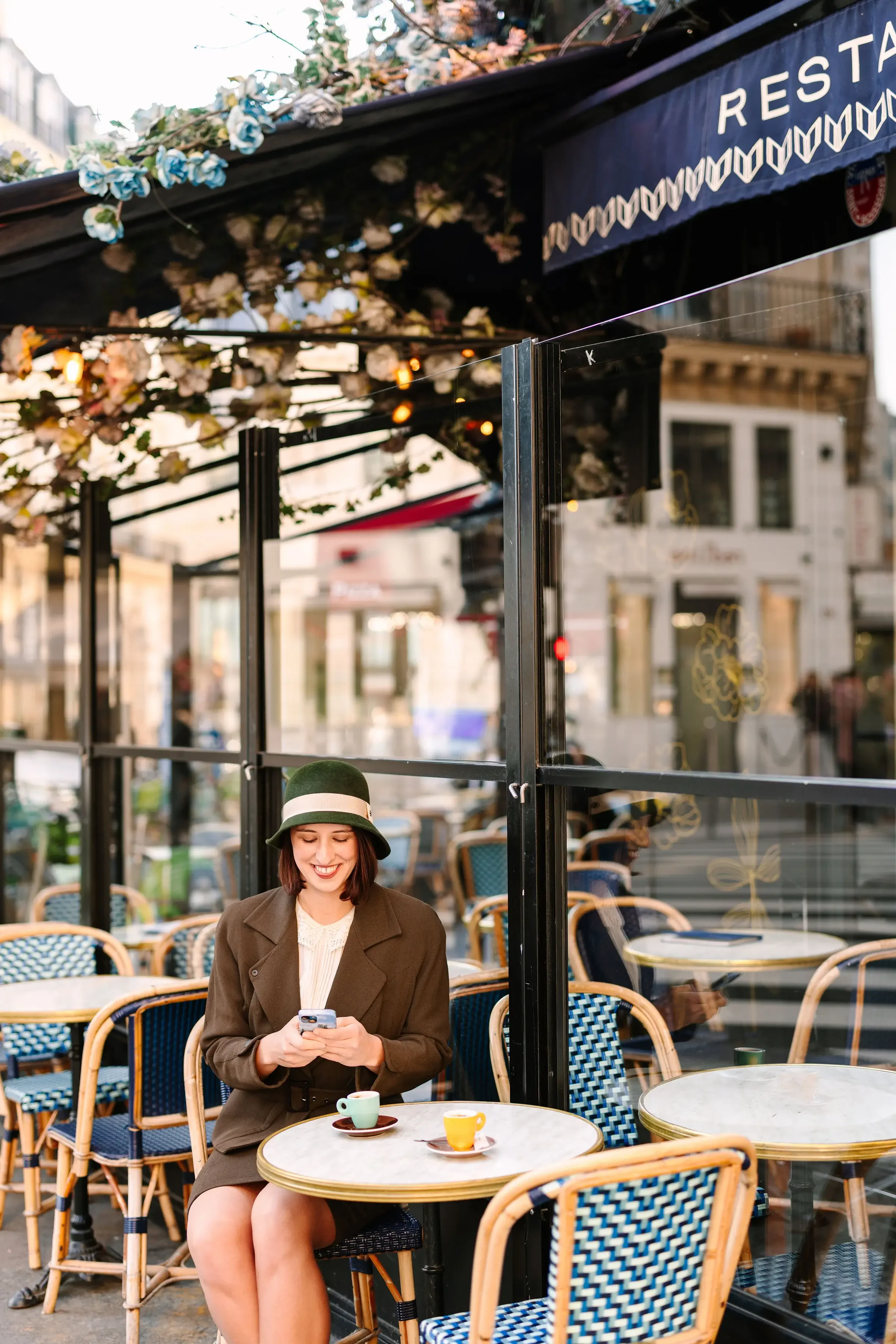 Hélène with shoulder-length brown hair wearing a dark blazer and a green hat with a white band, smiling while looking at her phone, sitting at an outdoor café with a cup of coffee or tea on a small round marble table.
