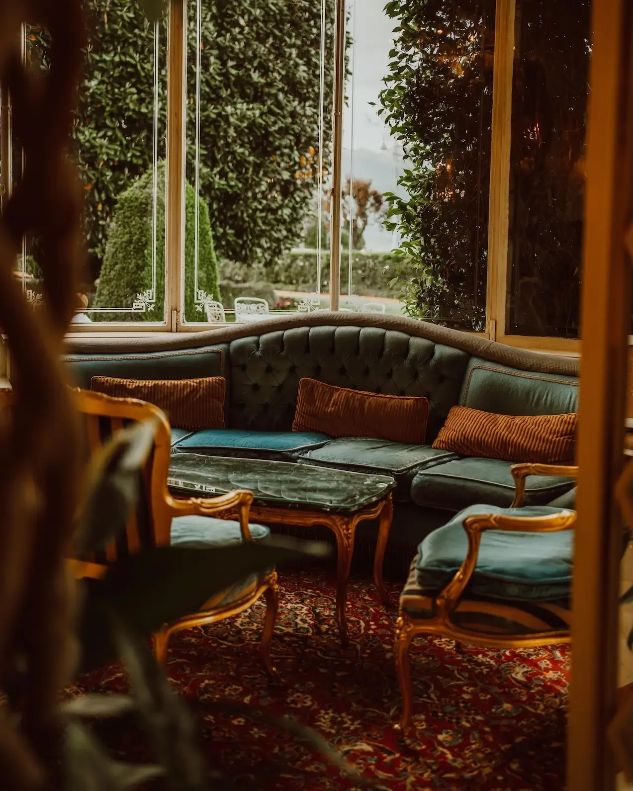 Vintage sitting area with a curved green velvet sofa, armchairs, and a marble-topped coffee table set on a patterned rug. Large windows reveal lush greenery outside.
