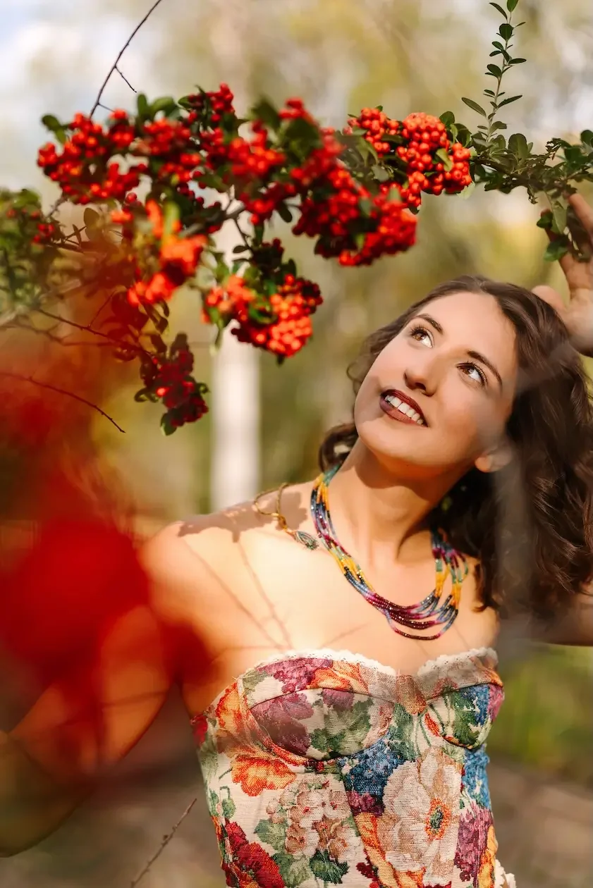 Hélène in a floral dress and multicolored beaded necklace poses outdoors near a branch with red berries while smiling and looking up.