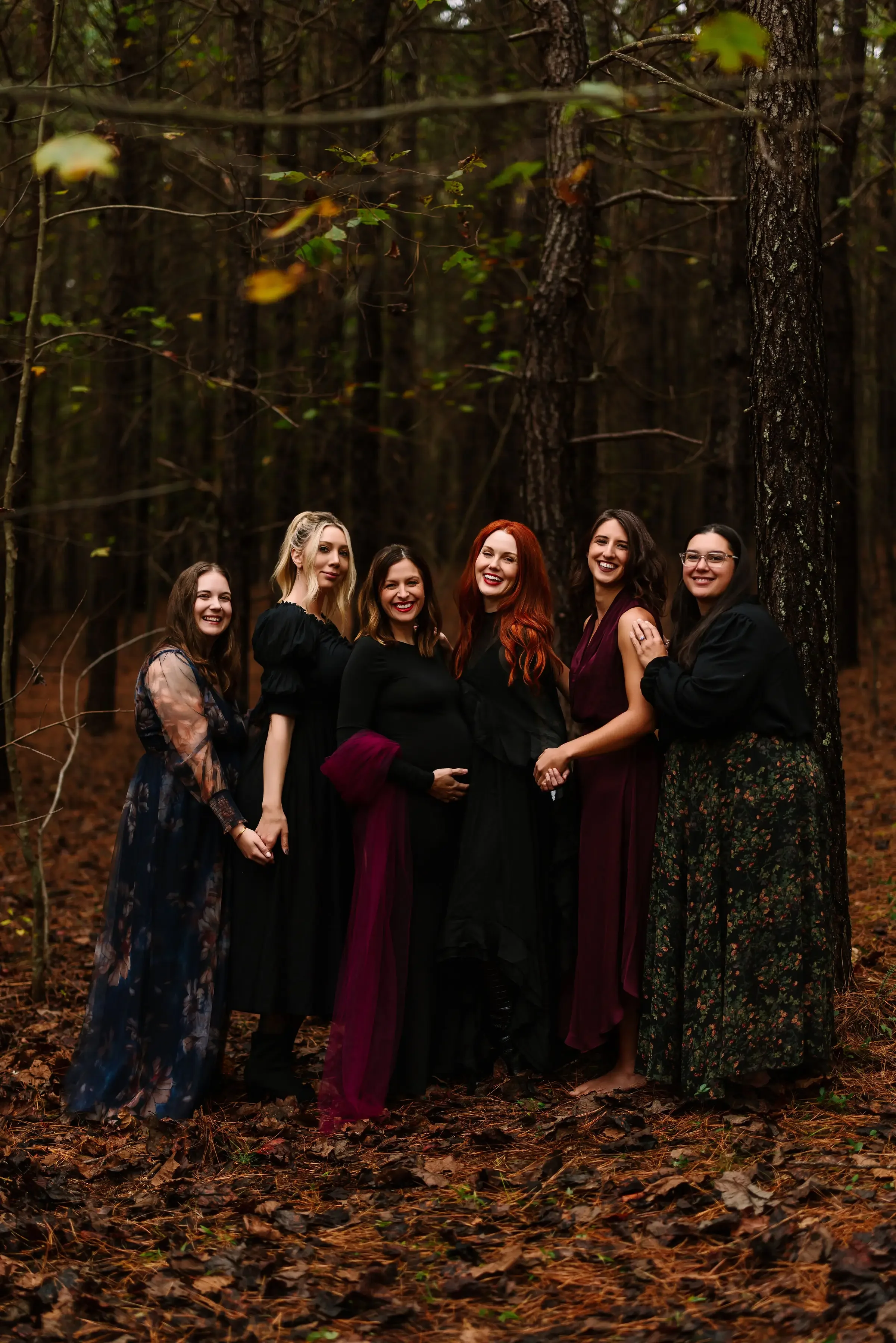 Group of seven women standing in a forest, dressed in dark and floral dresses, holding hands, with joyful expressions.