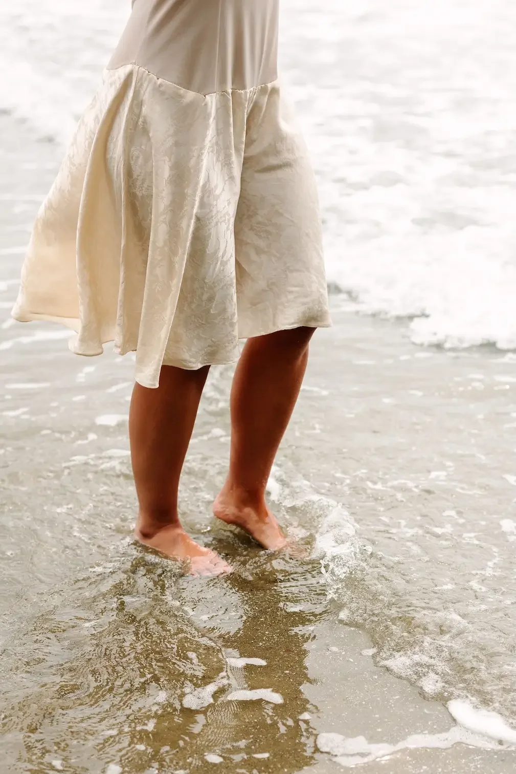 Hélène wearing a white dress is standing with her feet in the shallow water at the beach, with small waves around her ankles.