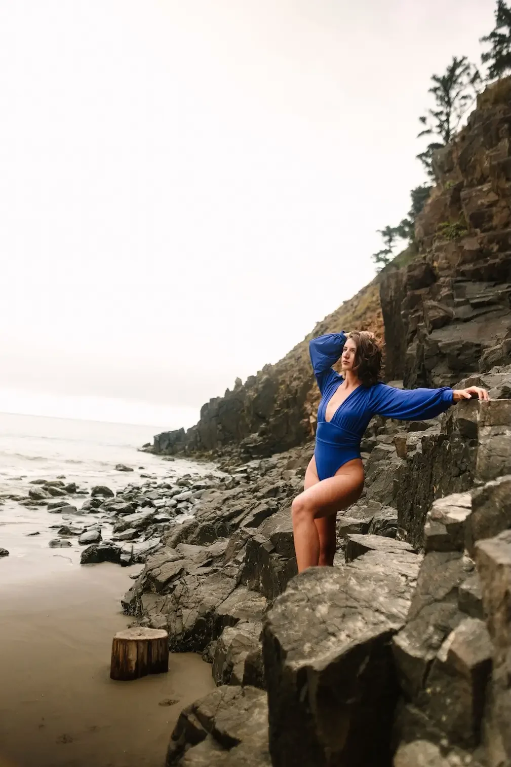 Hélène in a blue swimsuit standing on rocks by the ocean, holding onto a rock wall, with a cliff and trees in the background.