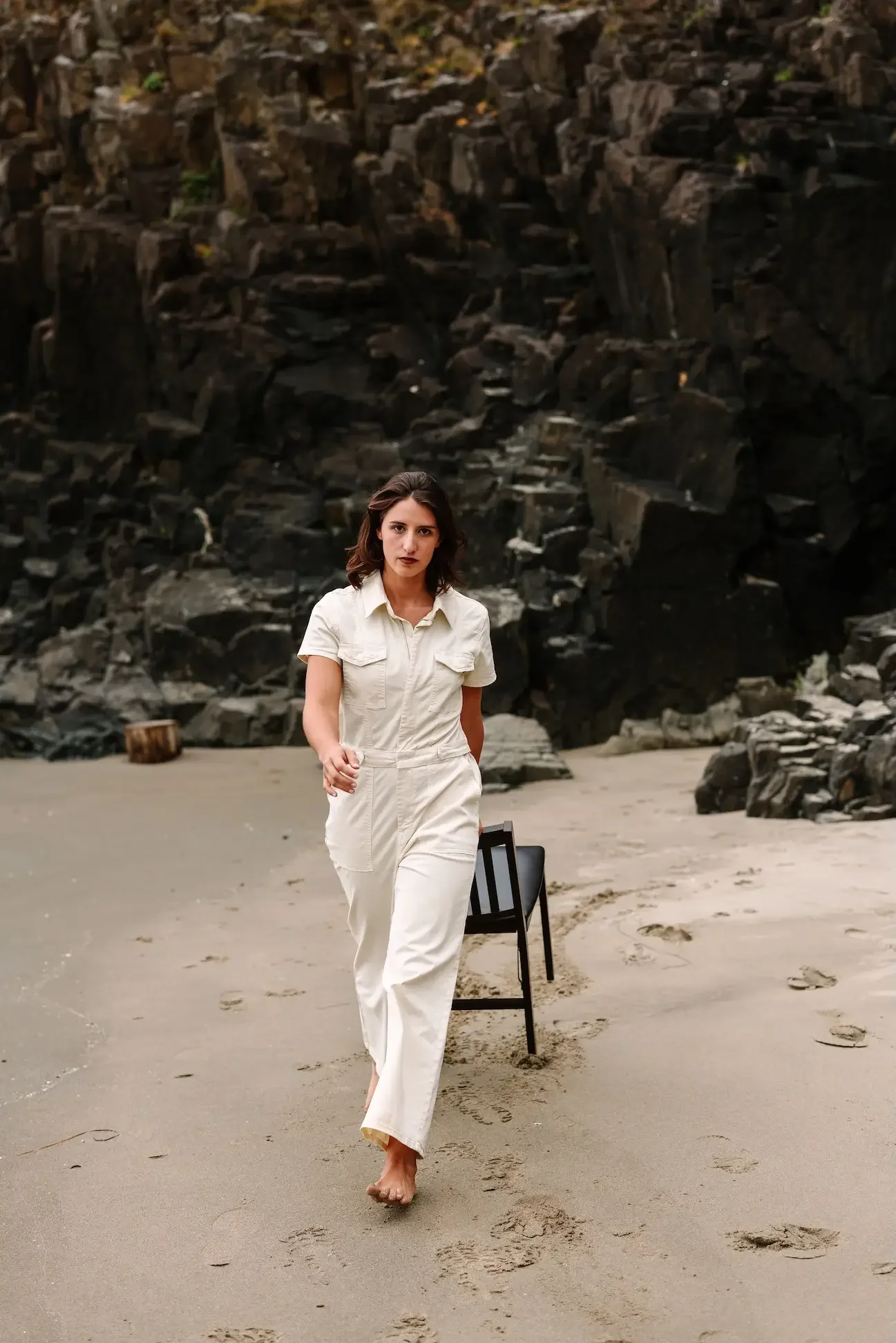 Hélène walking barefoot on a sandy beach with dark rock formations in the background, wearing a white jumpsuit.