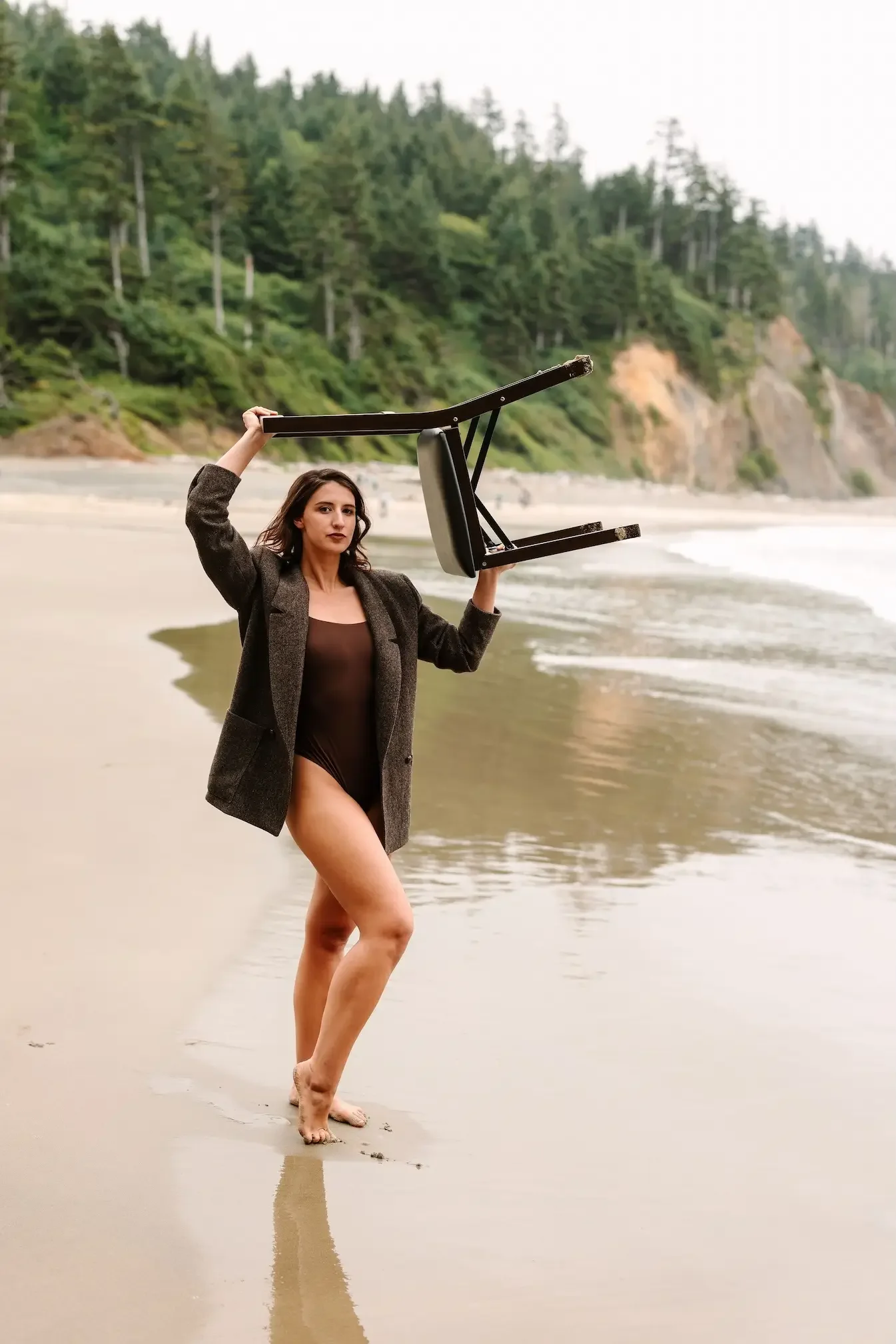 Hélène in a brown swimsuit and a brown blazer stands barefoot on a beach holding a chair above her head, with water and green trees in the background.