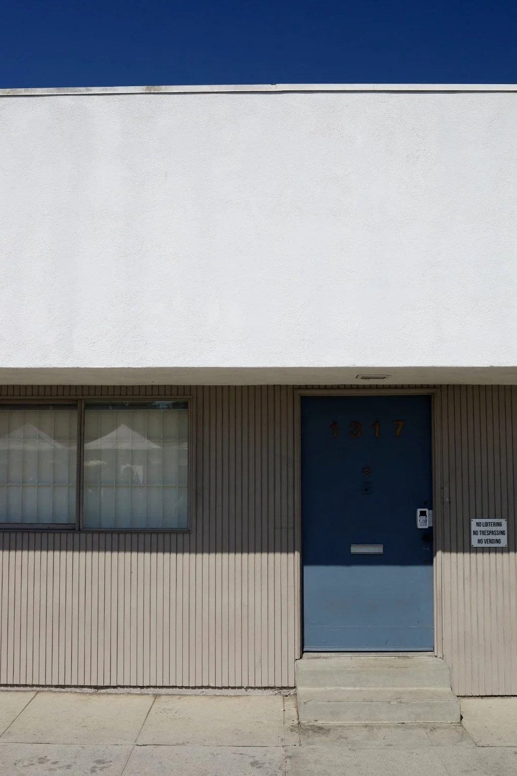 Ein schlichtes Gebäude mit blauer Tür, nummeriert 1917, und einem Fenster mit weißen Vorhängen. Es gibt eine Stufe vor der Tür und ein Schild mit den Hinweisen "No Loitering, No Trespassing, No Vending".