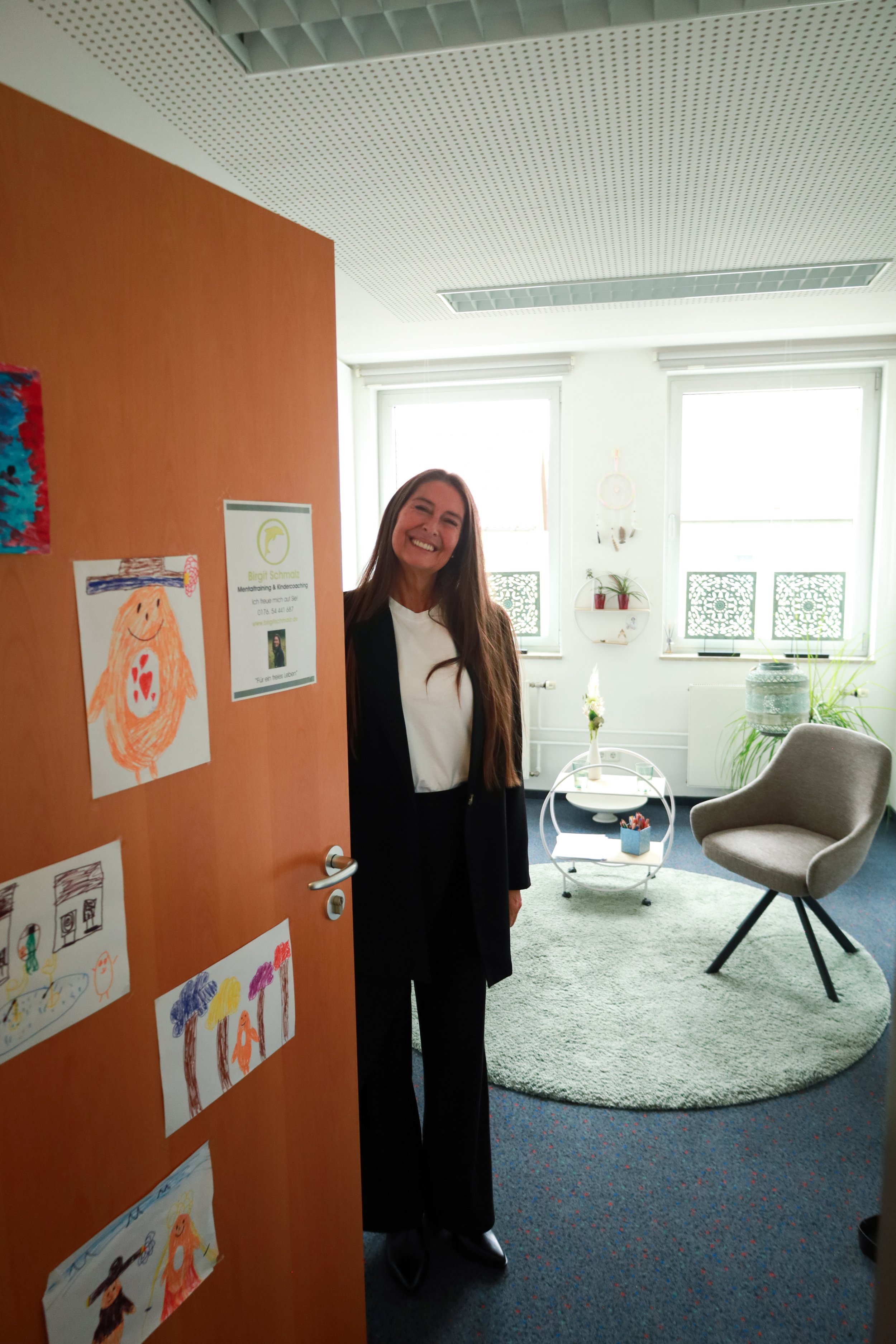 A woman with long brown hair, wearing a white top and black blazer, smiling and standing in a well-lit room with large windows, a gray armchair, and a circular green rug.