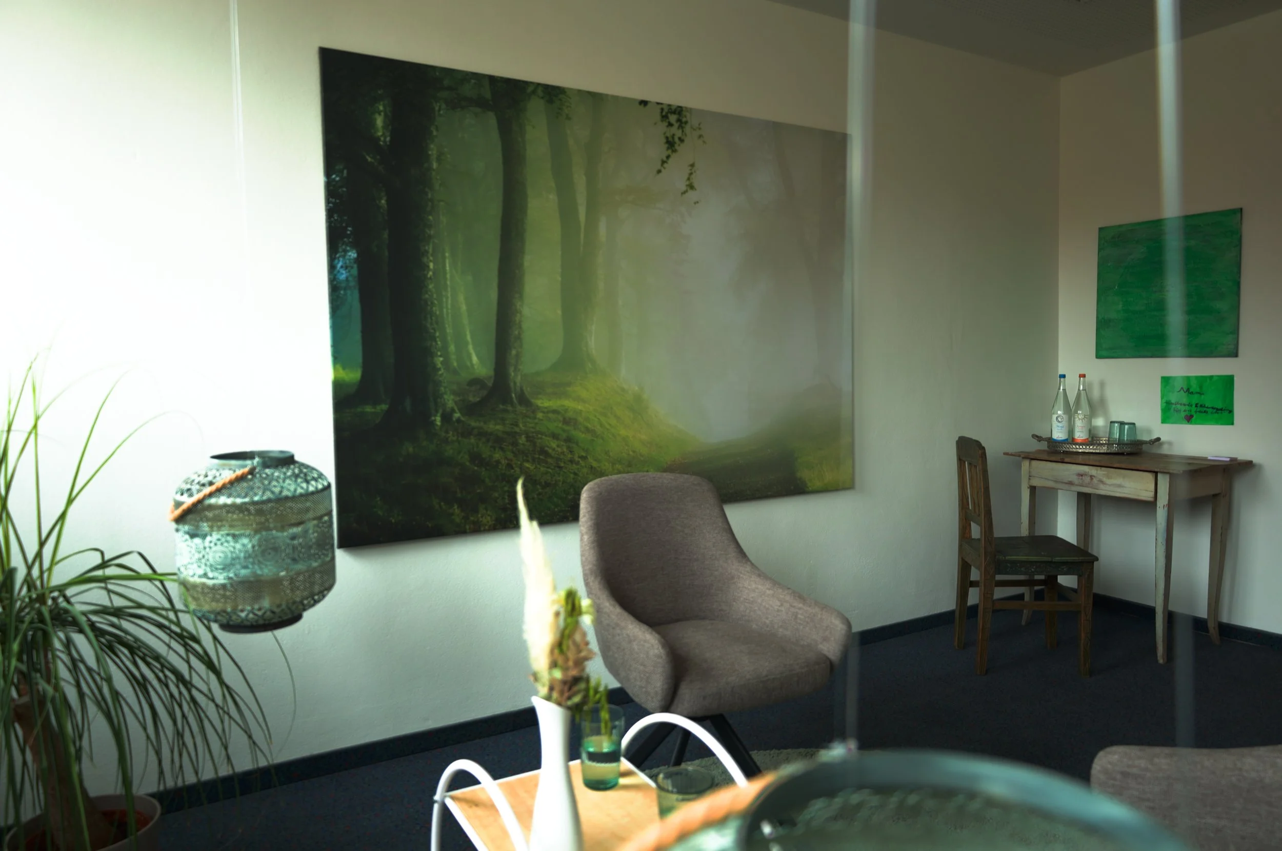 An interior view of a room with a large forest-themed painting on the wall, a gray armchair, a wooden table with bottles and glasses, and green artwork on the wall.