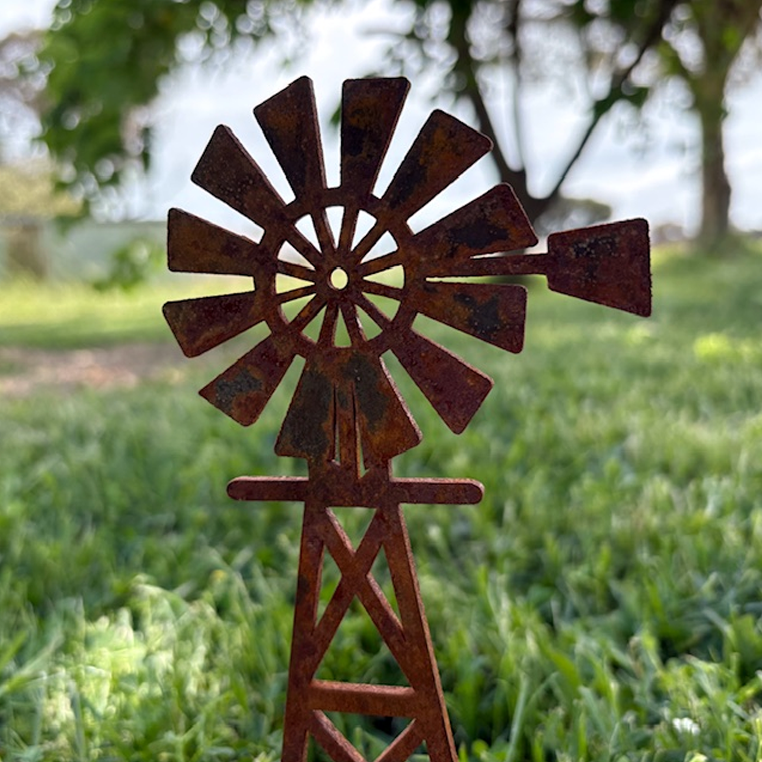 Close-up of a rusted metal windmill sculpture outdoors on green grass with trees and sky in the background.