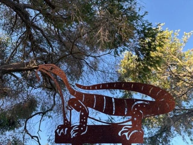 A rusted metal goanna lizard sculpture mounted with green leaves and a blue sky in the background.