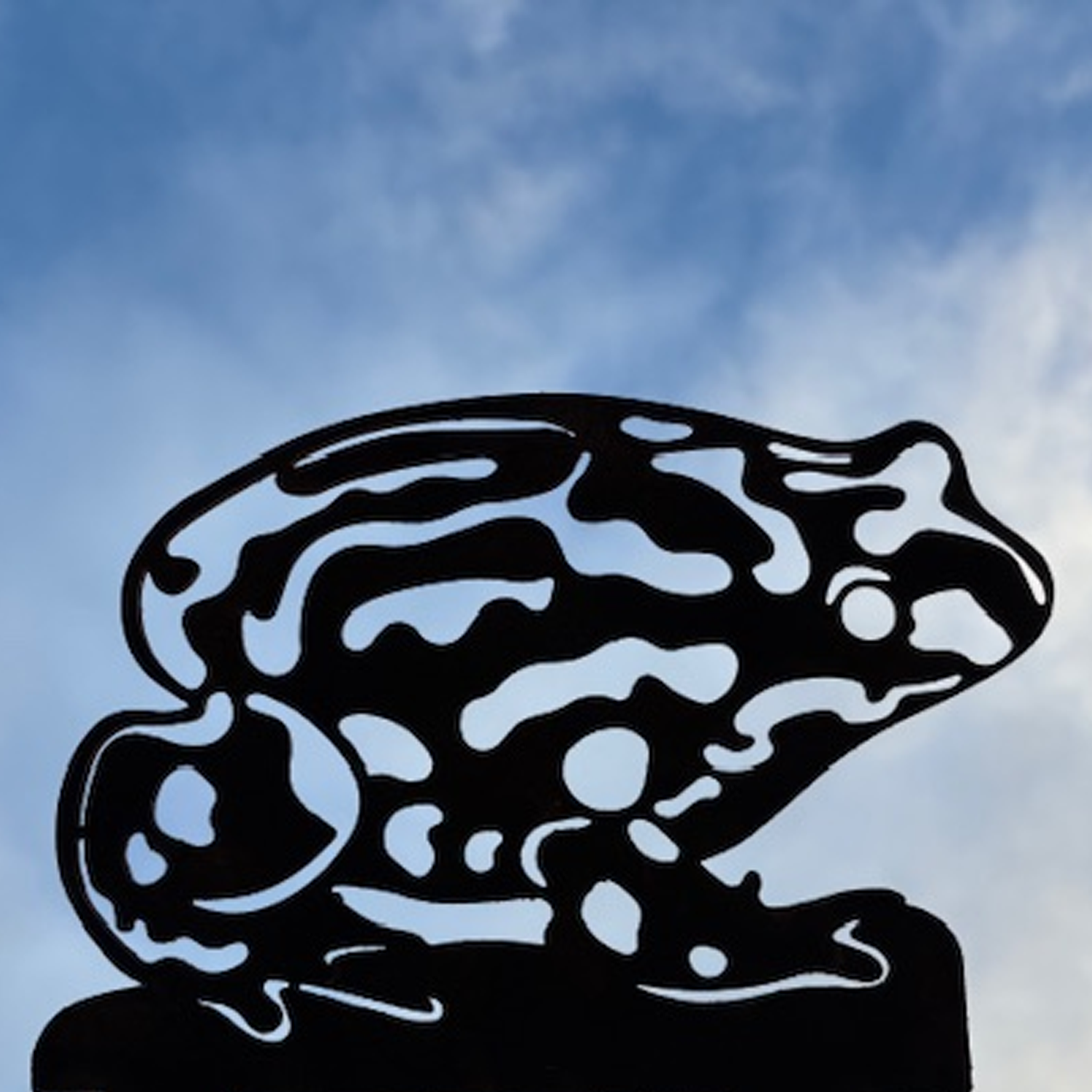 Black metal sculpture of a Corroboree frog with cut-out patterns against a cloudy sky background.