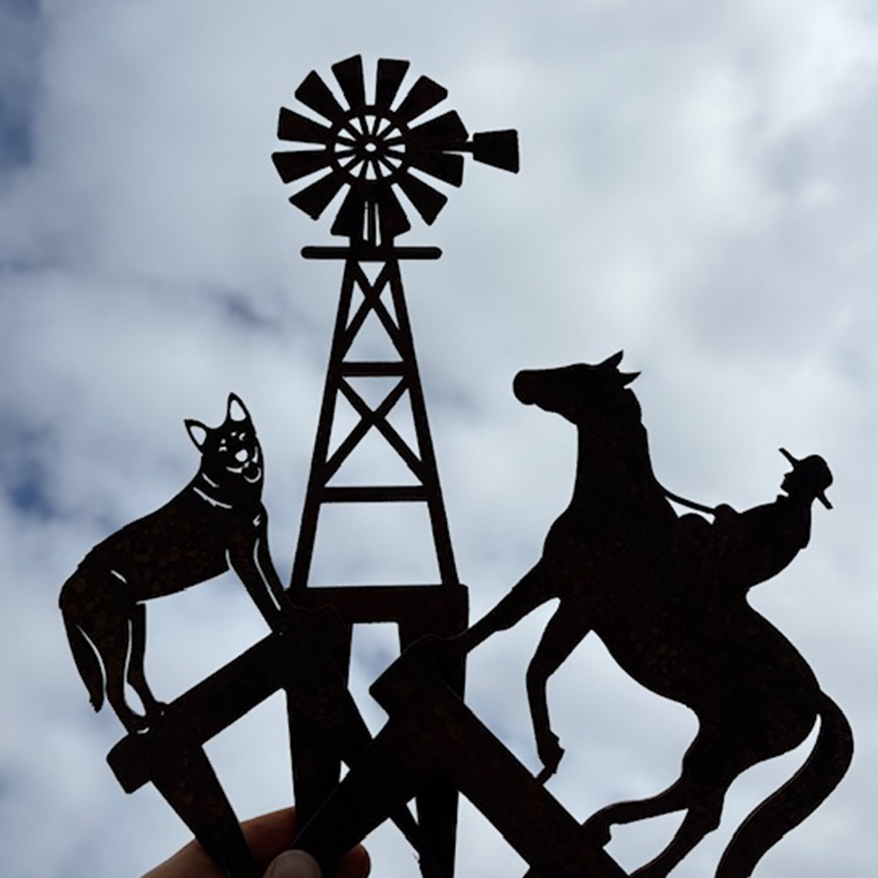 Silhouette of metal sculptures featuring a Blue Heeler dog, a cattle hand on a horse, and an Outback windmill against a cloudy sky.