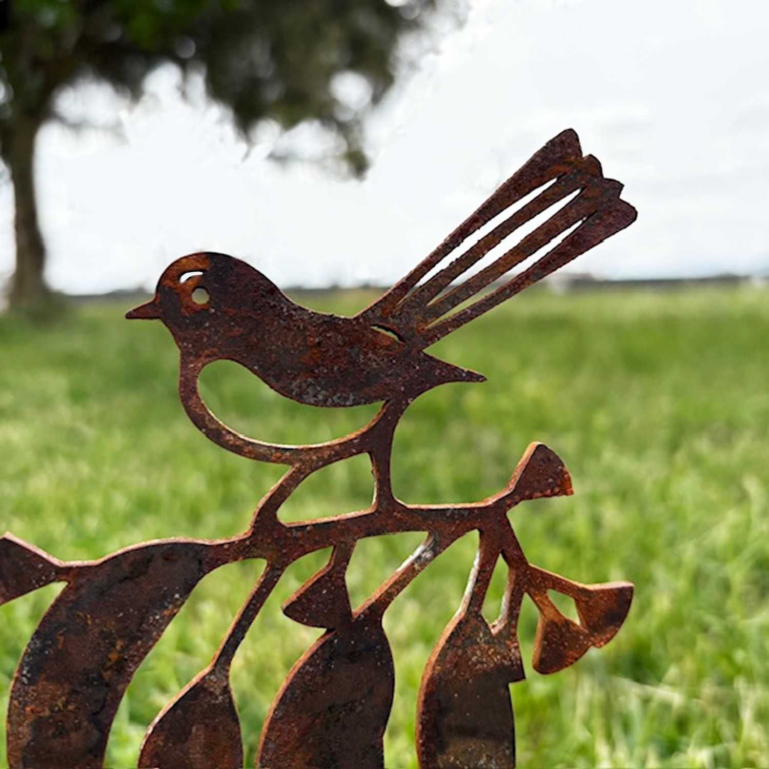Rusty metal sculpture of a Willy Wagtail bird perched on a branch, with a grassy field and trees in the background.