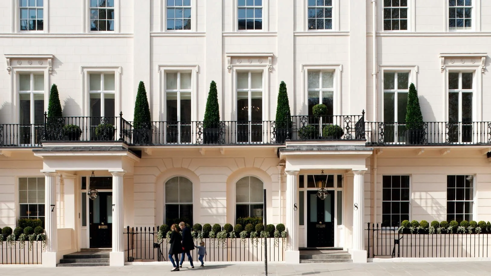 White multi-story building with black doors, decorative columns, potted topiary, and black wrought iron balcony railing; three people walking in front.