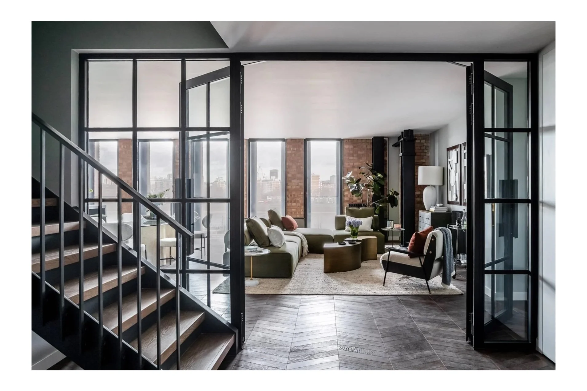 Modern urban living room with large windows, contemporary furniture, a staircase, and city skyline views.