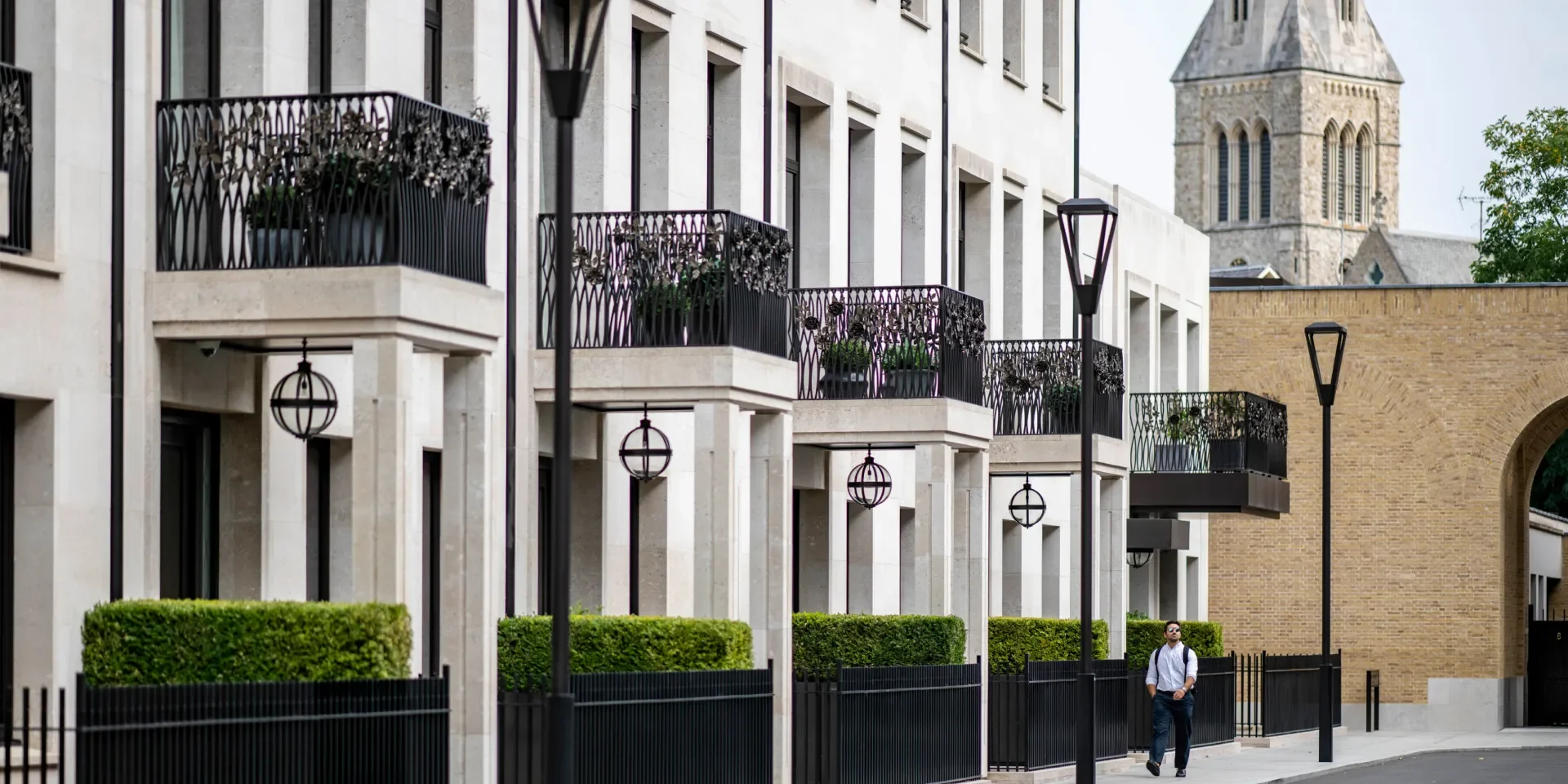 Urban street scene featuring modern apartment buildings with balconies, decorative black fencing, and classical church tower in background. Pedestrian with backpack walking on sidewalk, black street lamps, well-maintained green bushes in foreground.