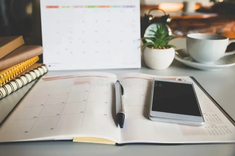 Open planner with a pen, smartphone, and various notebooks on a desk. In the background, there are a potted plant, a coffee cup on a saucer, and a blurry computer monitor.