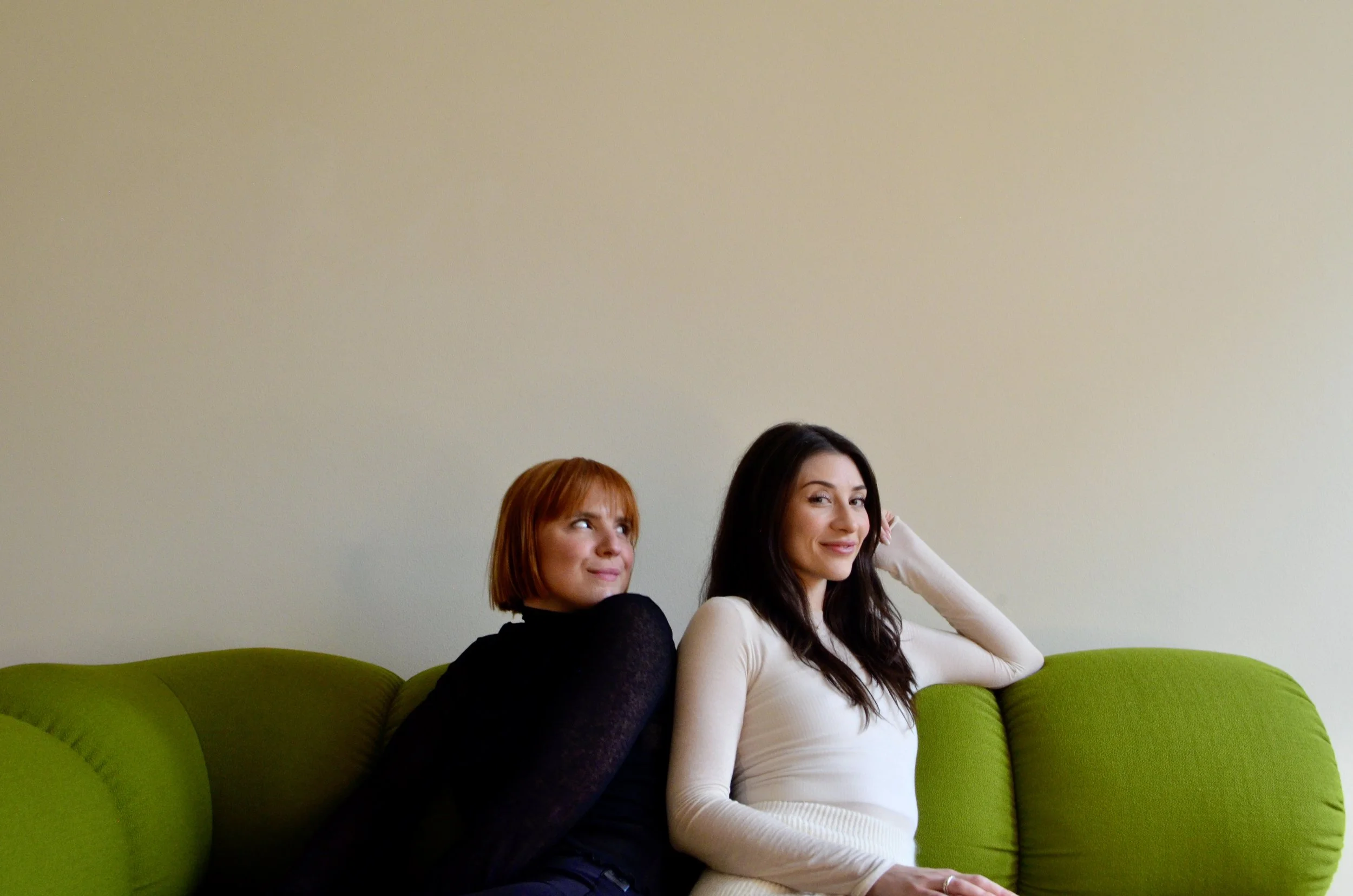 Two women sitting on a green sofa against a beige wall, one with red hair wearing a black top, the other with dark brown hair in a white top, smiling and looking at each other.