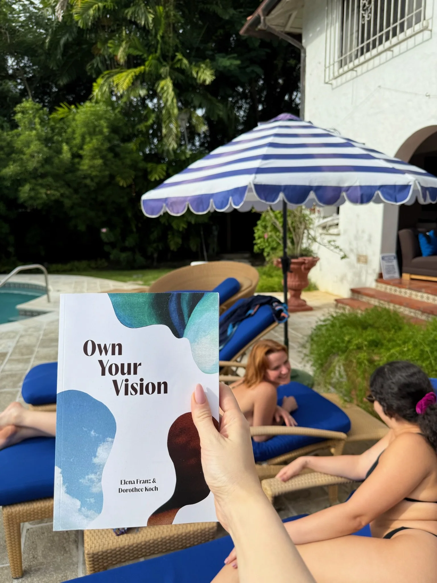 A person holding a book titled 'Own Your Vision' by Elena Franz & Dorothee Koch in a backyard with a swimming pool, lounge chairs, a blue and white striped umbrella, and people relaxing by the pool.