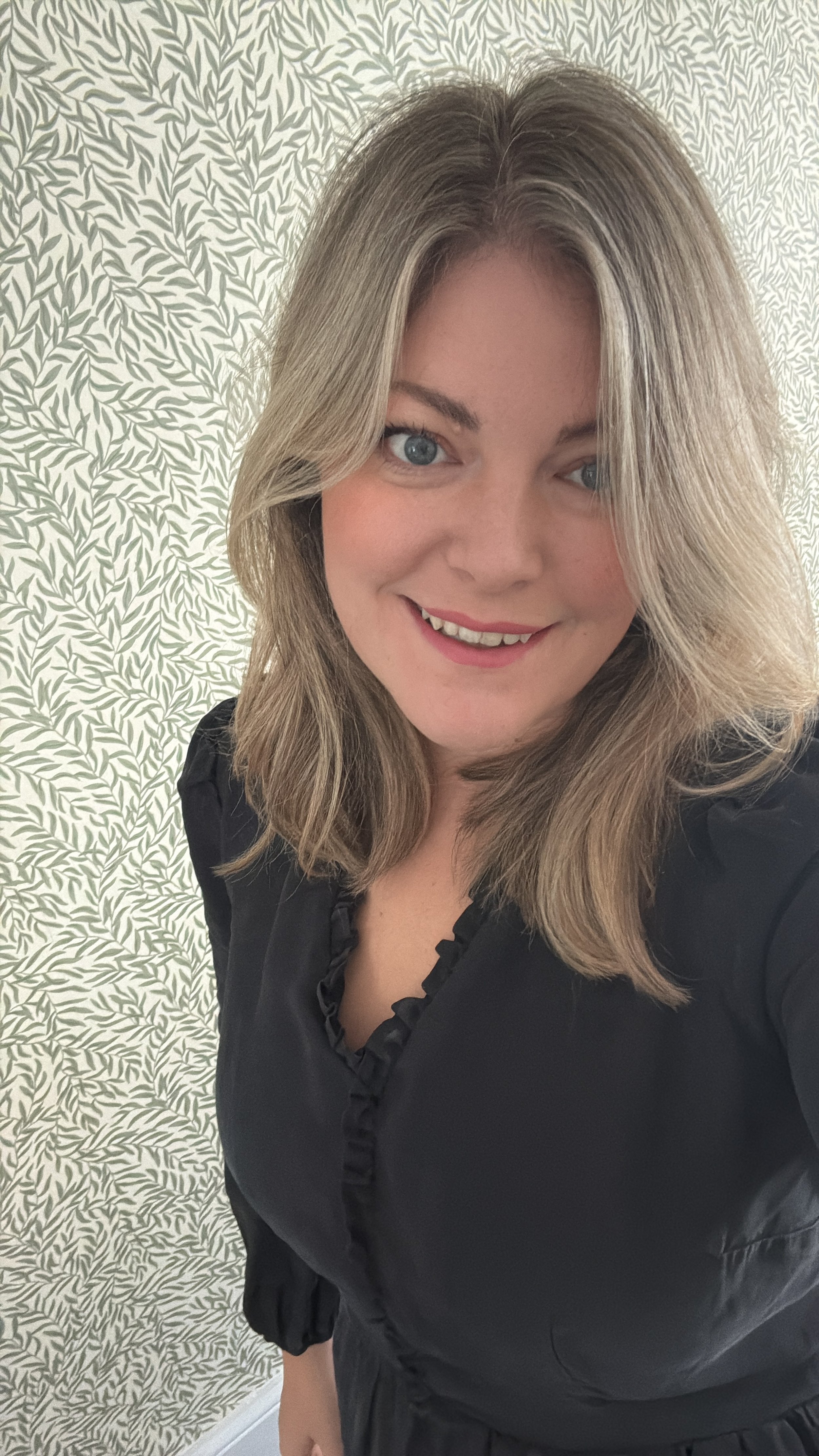 Headshot of Jo Pope. Blonde hair and blue eyes, taken against a leafy wallpaper background, wearing a black blouse.