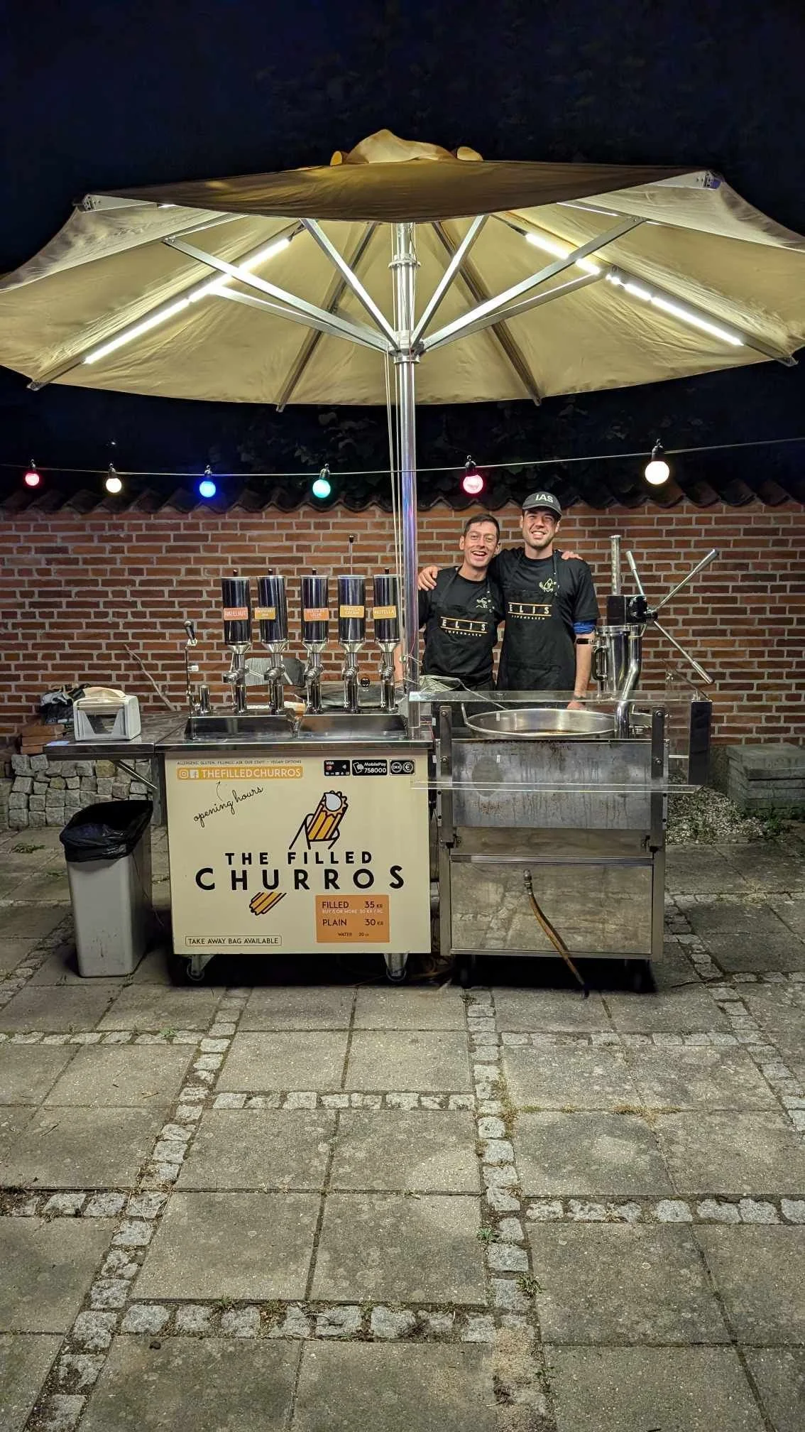 Two men standing behind a soft serve ice cream kiosk named 'The Filled Churros' under a yellow umbrella at night. The kiosk has multiple dispensers and a sign with prices for filled and plain churros. The background features a brick wall and multicolored string lights.