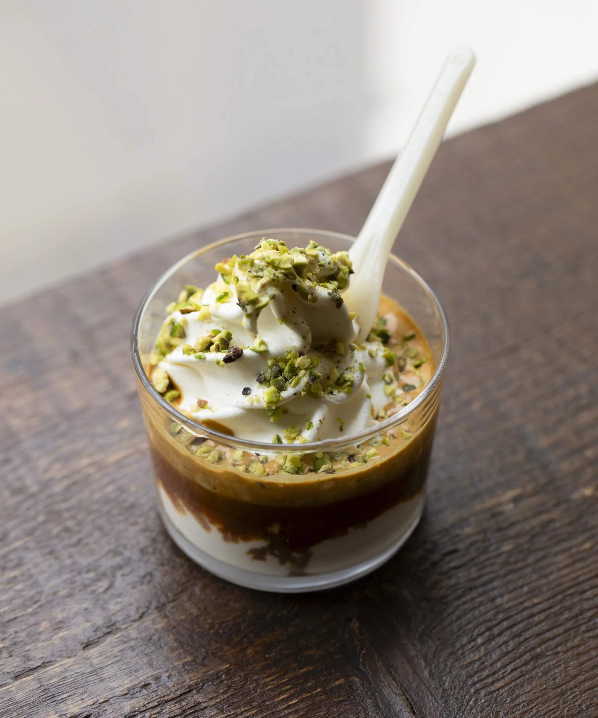 Creamy dessert in a glass cup with whipped cream, chopped pistachios on top, and a white spoon, on a wooden surface.
