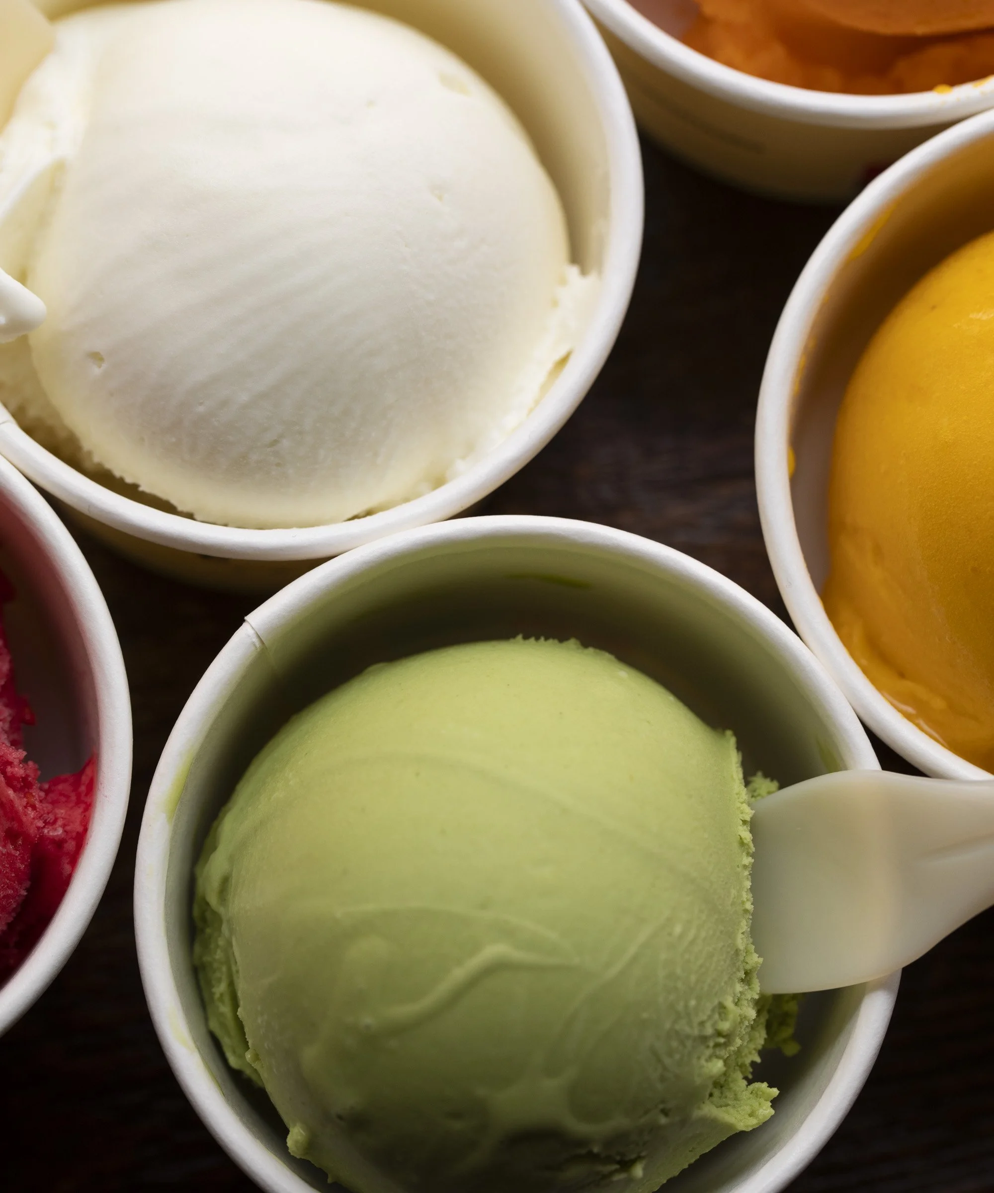 Multiple cups of ice cream in different flavors, including vanilla, pistachio, and mango.