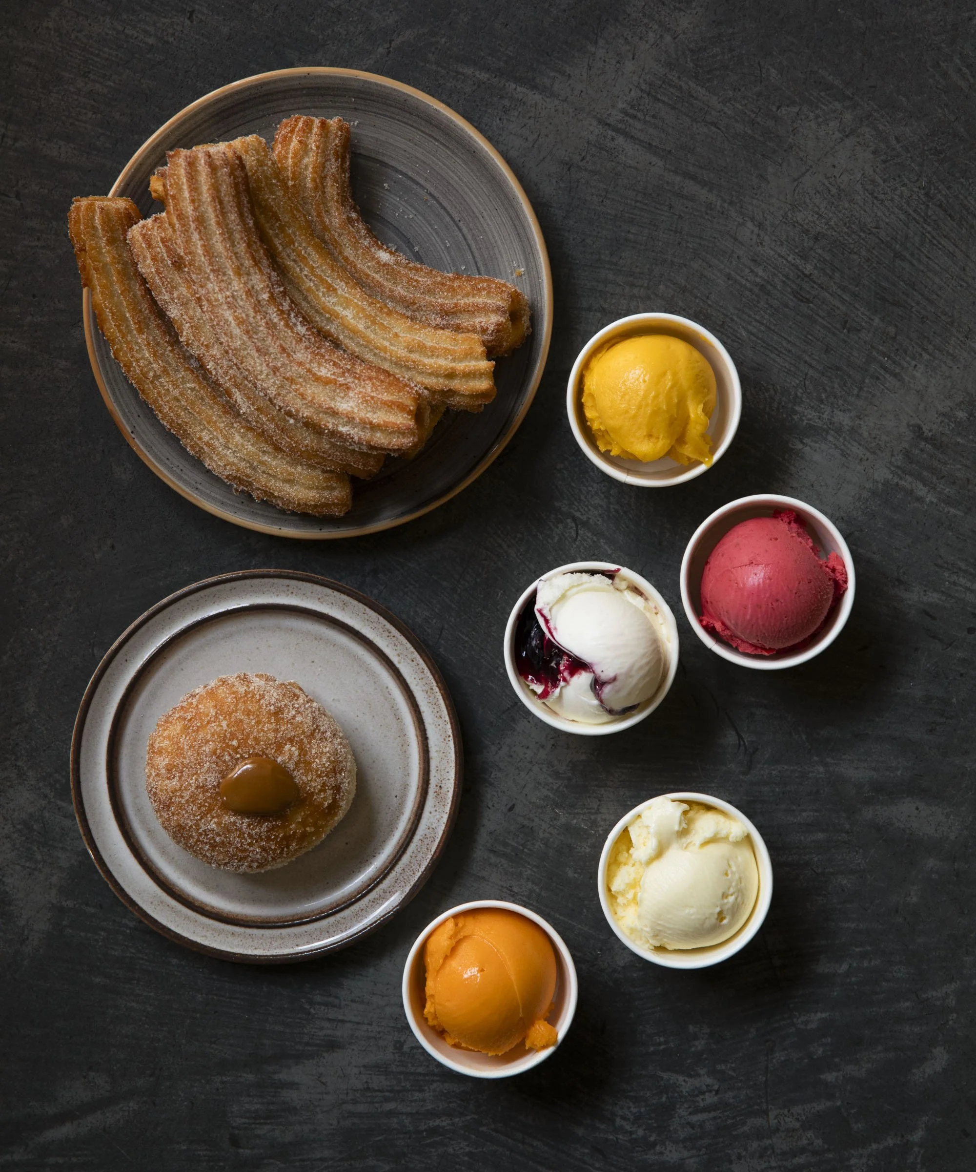 Churros with sugar served on a plate, surrounded by six small cups of different flavored ice creams.