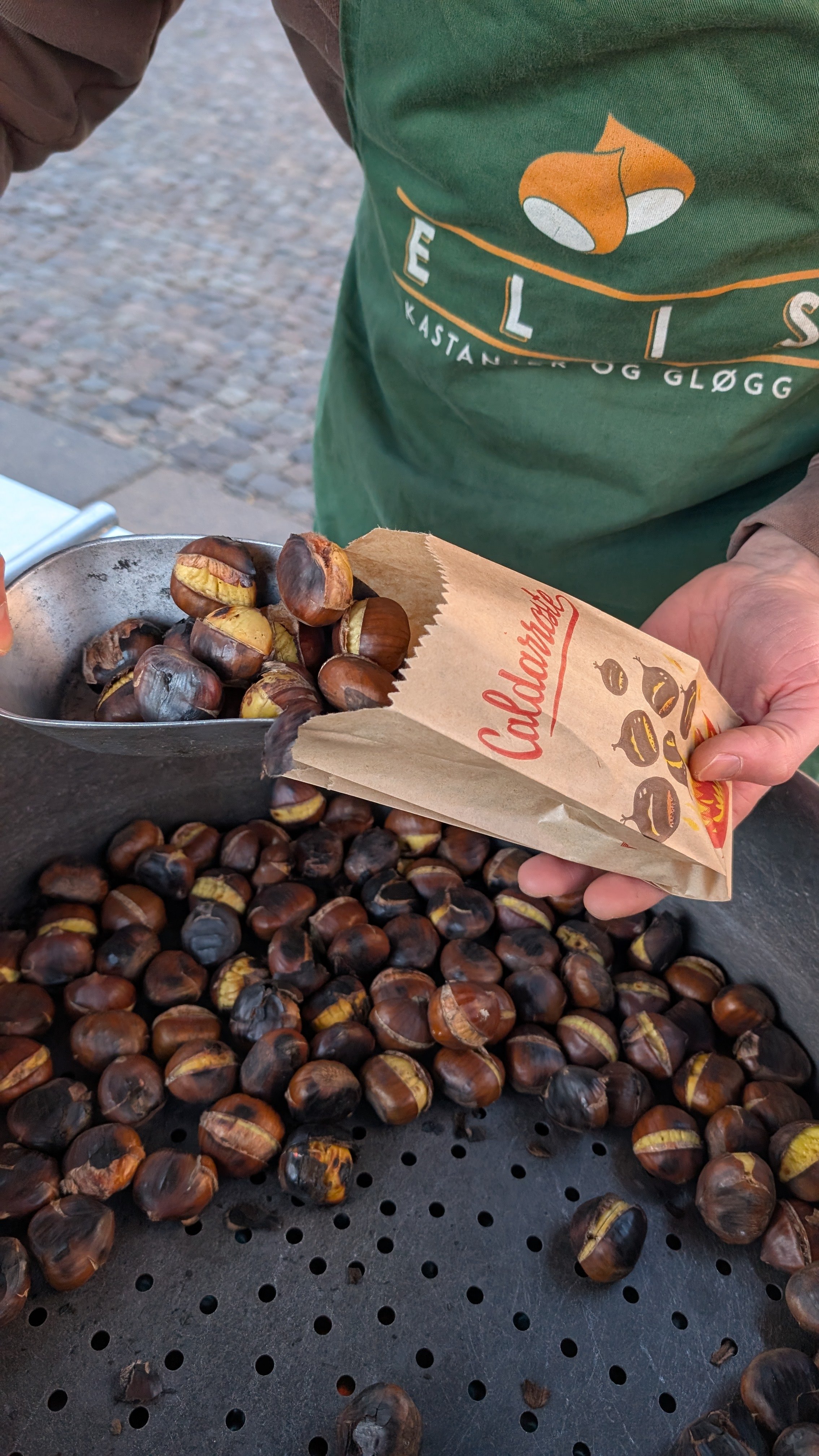 Person holding a paper bag of roasted chestnuts over a large tray of roasted chestnuts on a black perforated surface outdoors.
