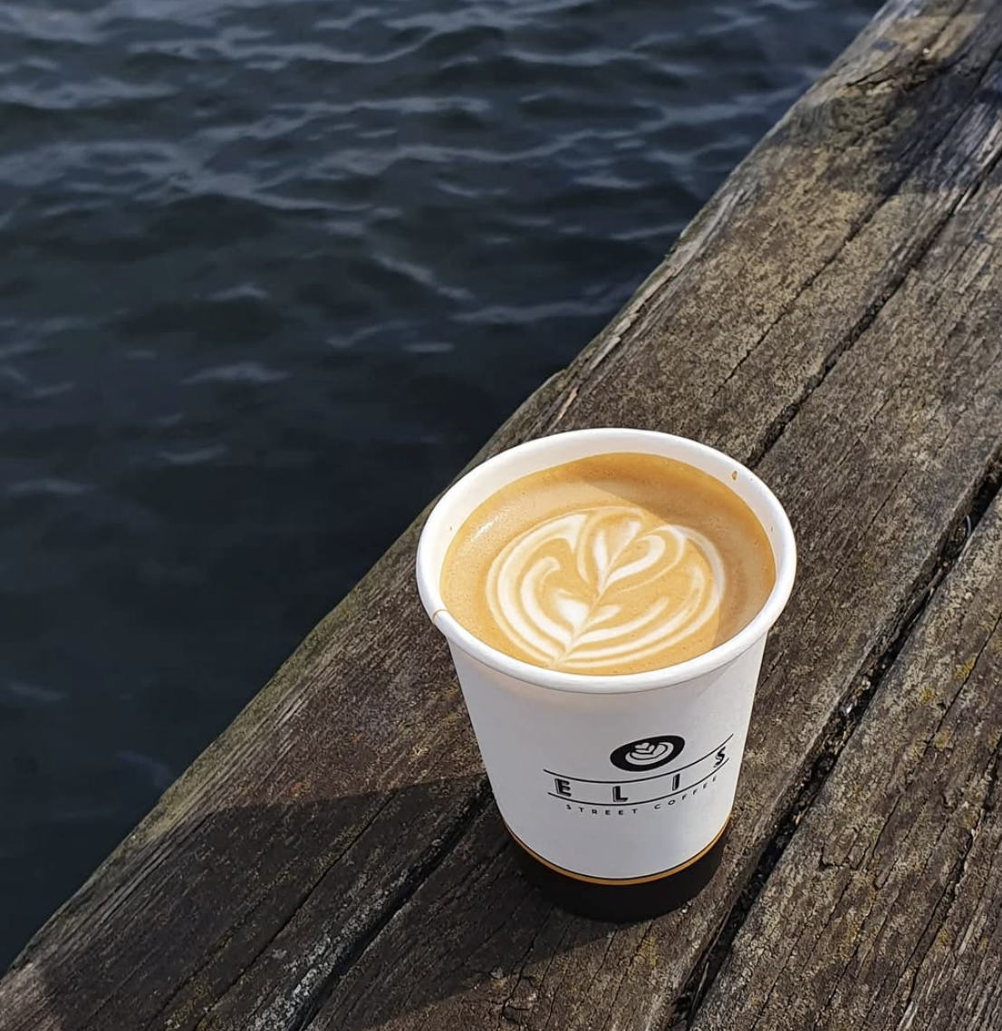 A cup of coffee with latte art on a wooden dock over water.