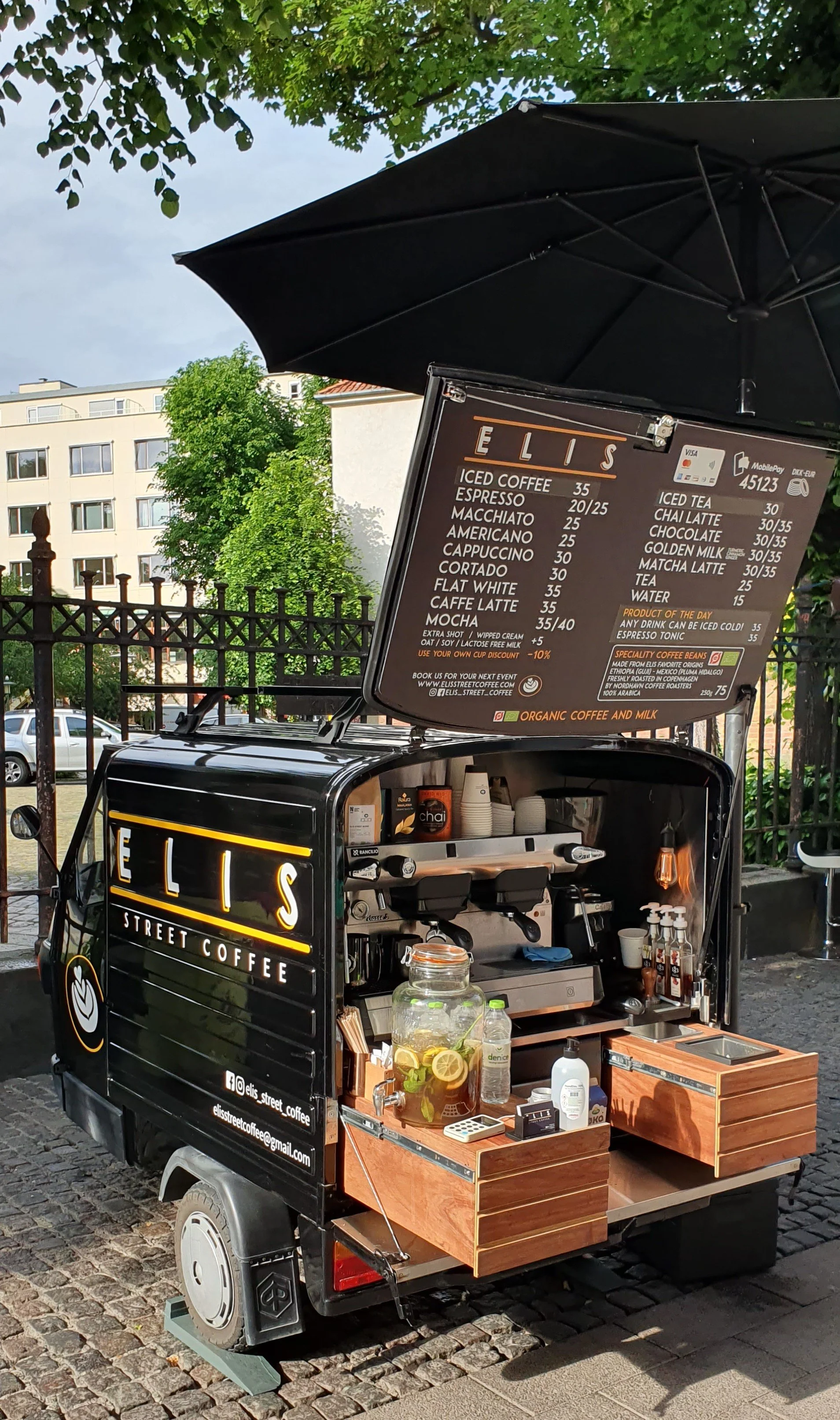 A black mobile coffee stand with a menu board on top, selling various coffee drinks and beverages. The stand is outdoors on cobblestone pavement with trees and buildings in the background.