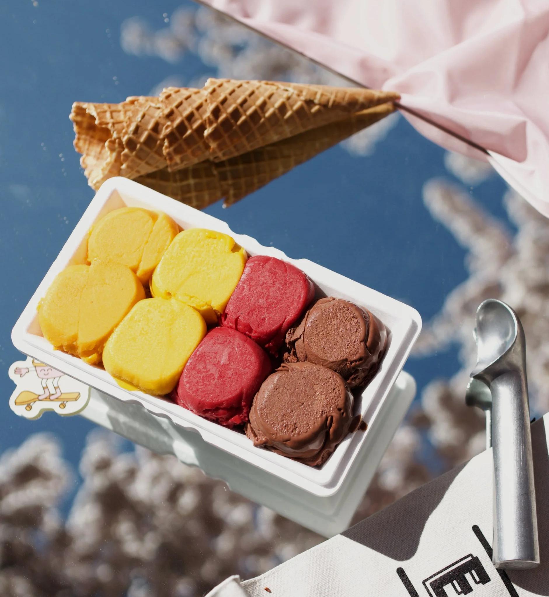 A white box of assorted ice cream scoops, including yellow, pink, and brown flavors, with a blurry background.
