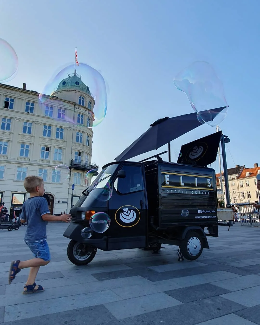A small street coffee vehicle with the signage 'ELIS Street Coffee' is parked on a city street, with a child running past it and bubbles floating in the air. The background features historic-style buildings and a domed tower under a clear blue sky.