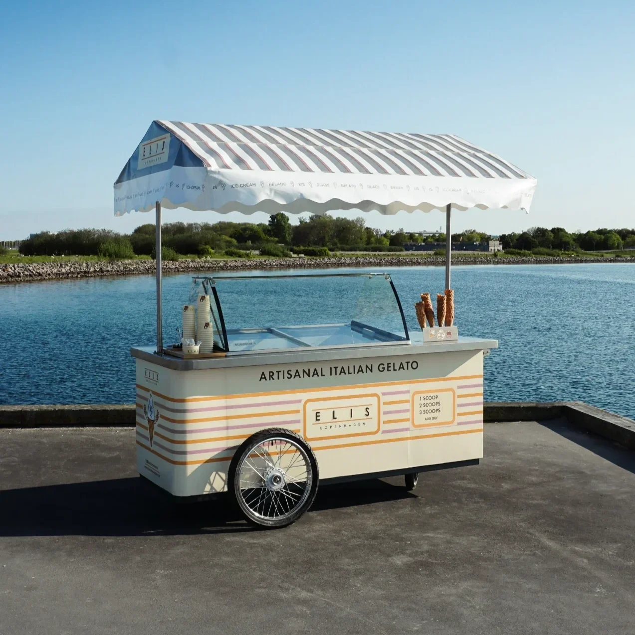 Ice cream cart on pavement near water with green trees and blue sky in the background. The cart has a striped umbrella and is labeled "Artisanal Italian Gelato."