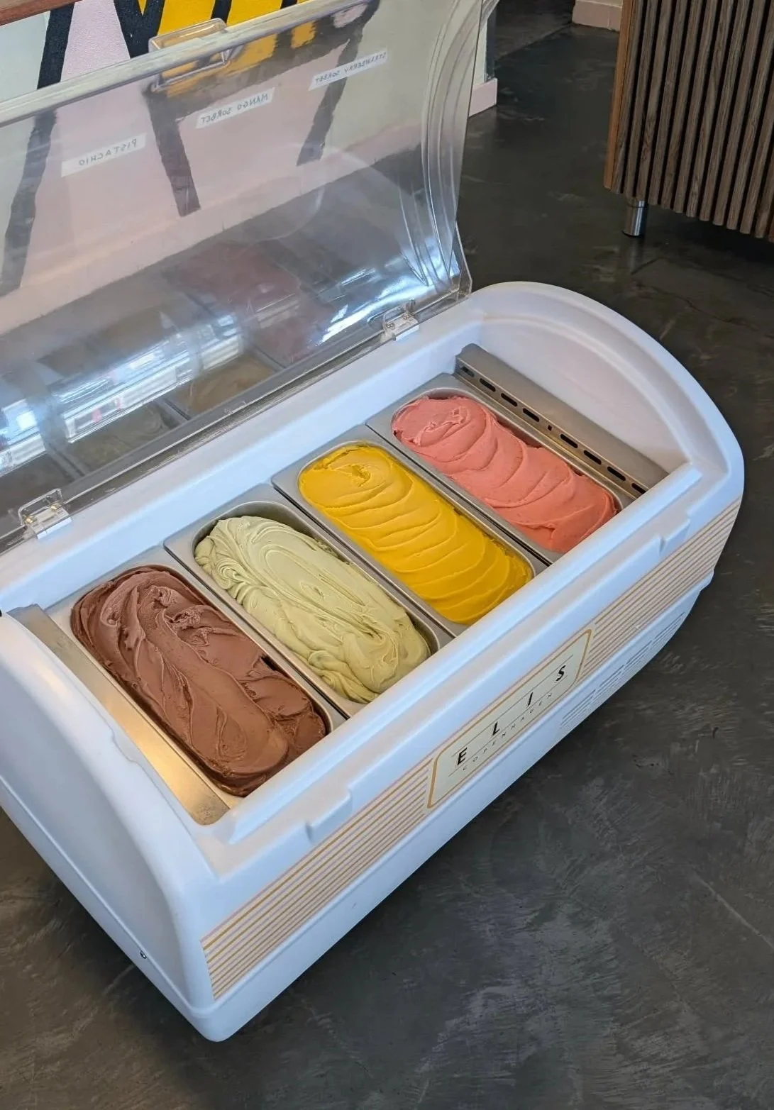 A white ice cream freezer with a transparent lid, containing four flavors of ice cream in metal containers. From left to right: chocolate, vanilla, mango, and strawberry ice cream. The freezer is on a dark gray floor, with part of a wooden lattice visible on the right side.