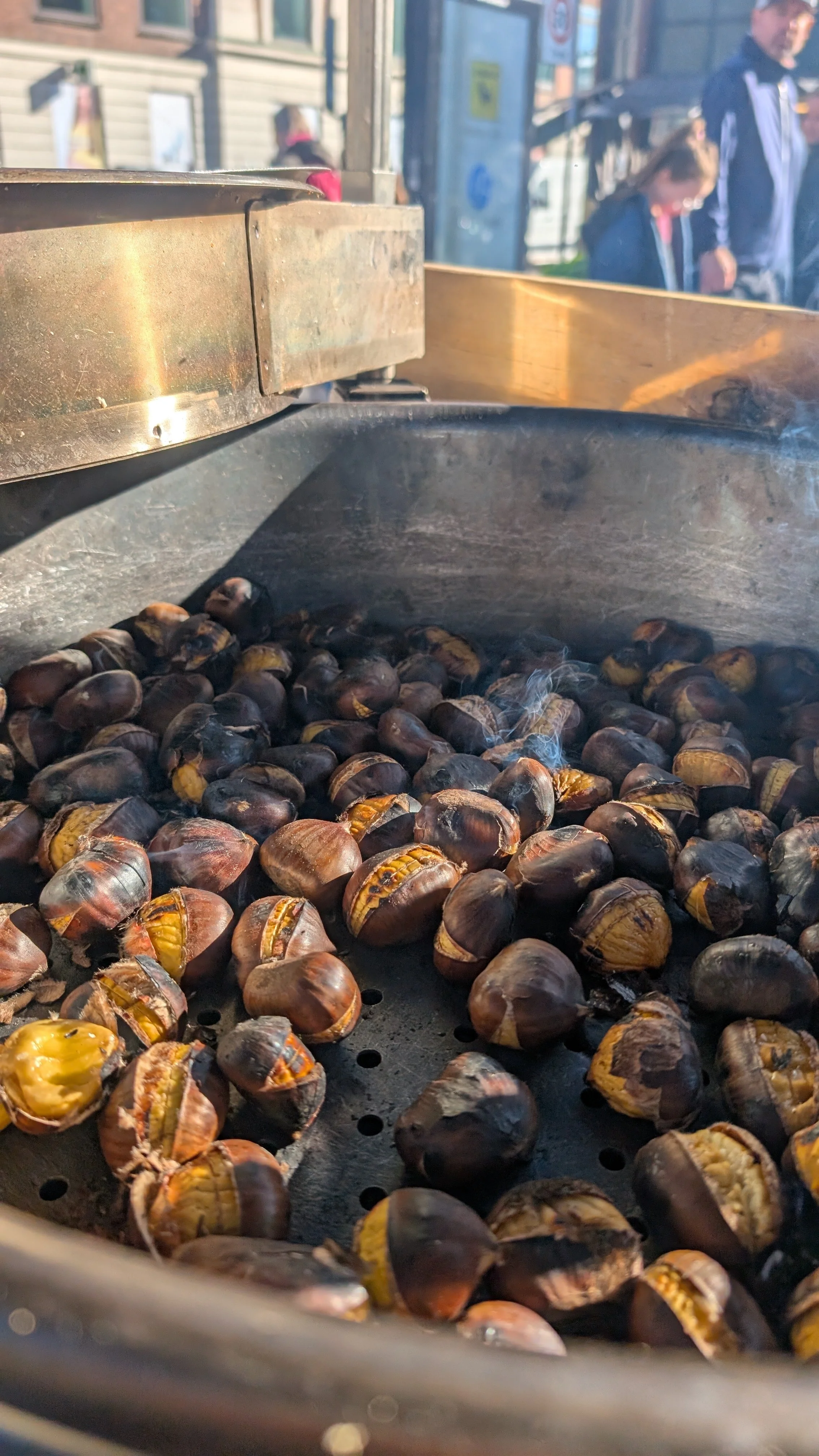 Roasted chestnuts in a pan with some smoke, outdoors on a sunny day with people walking in the background.