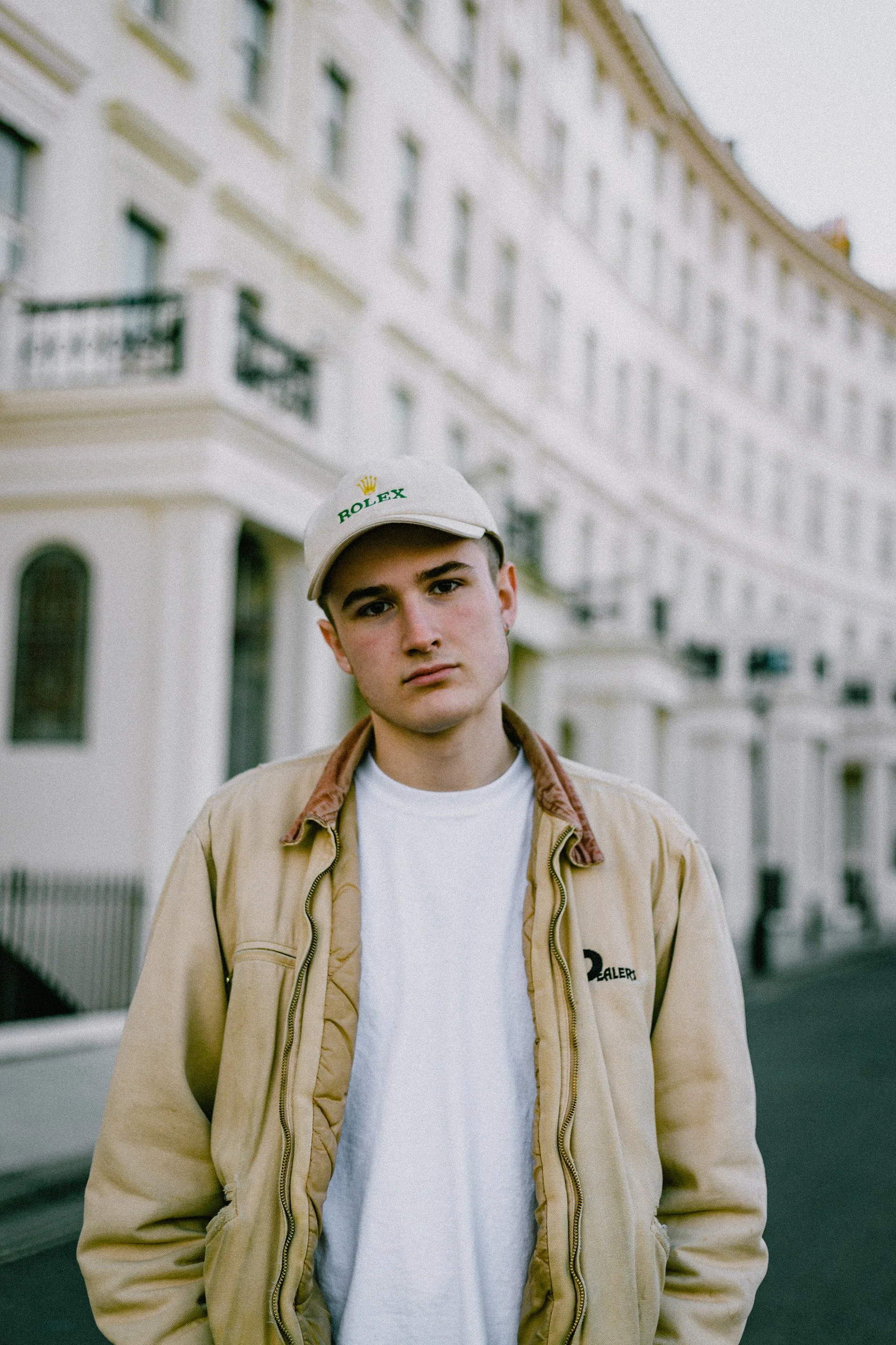 Young man Zach wearing a beige jacket and a white cap with 'ROLEX' logo, standing on a city street with white multi-story buildings in the background.