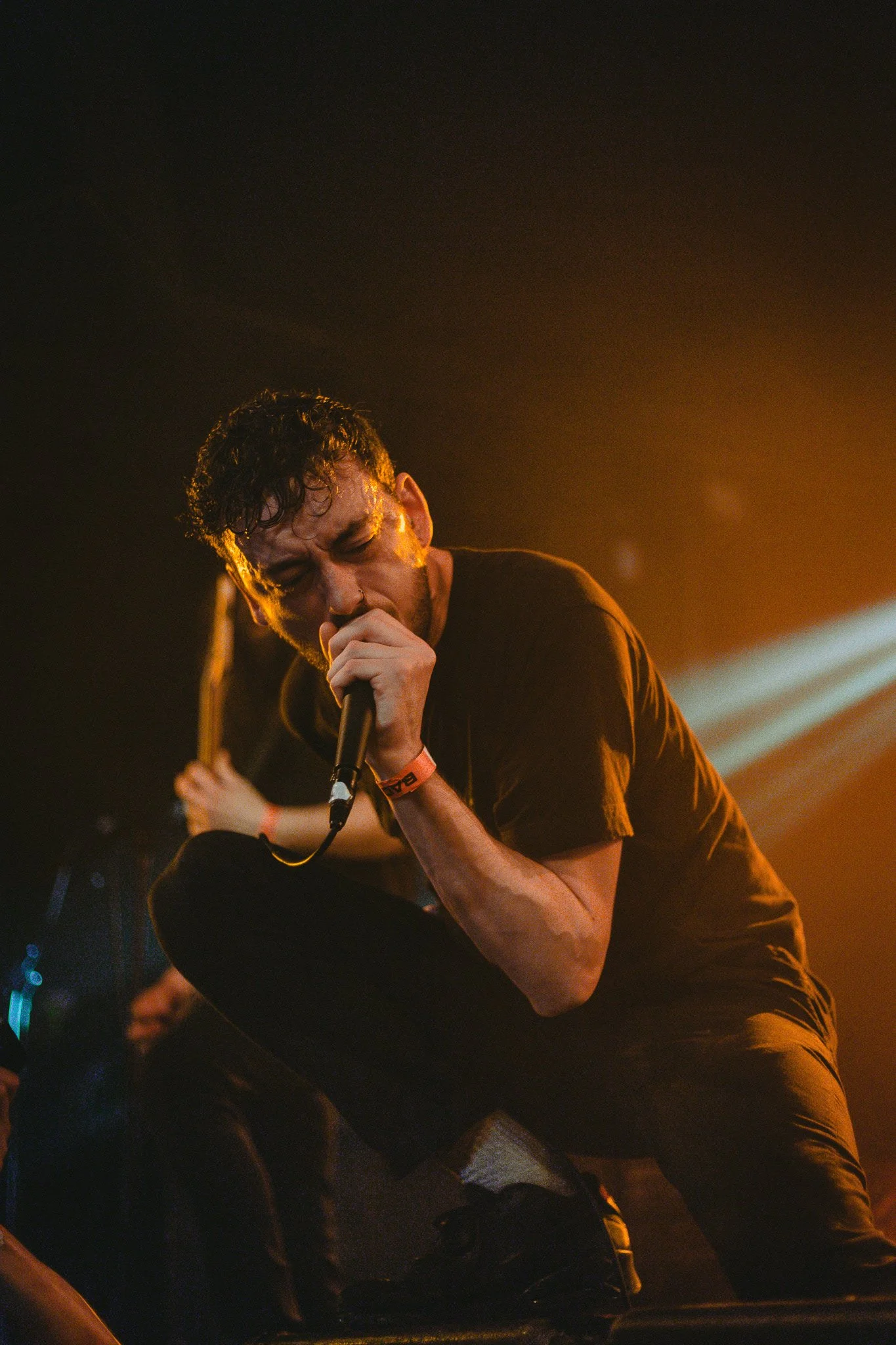 Male singer with curly hair and black t-shirt, kneeling and holding a microphone on stage, illuminated by stage lights.