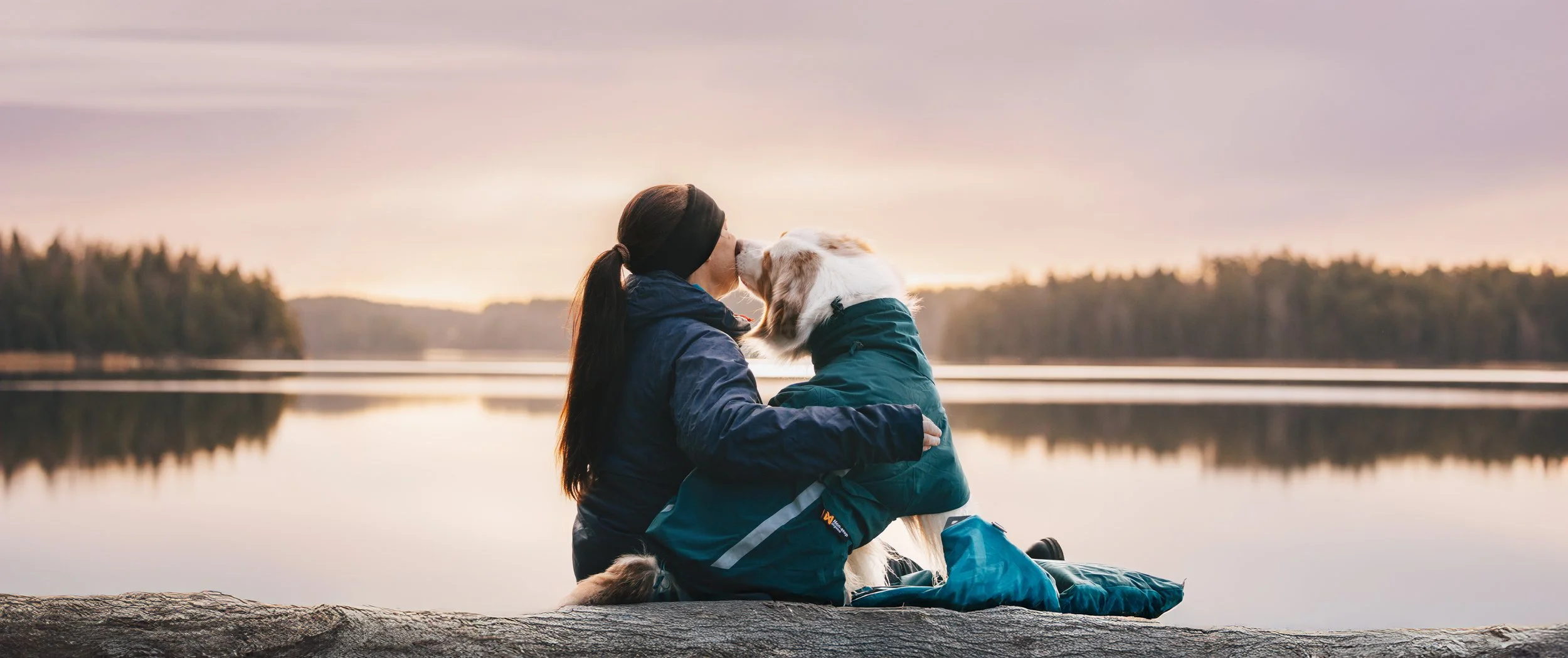 A person and a dog sitting on a log by a calm lake at sunset, sharing a kiss.