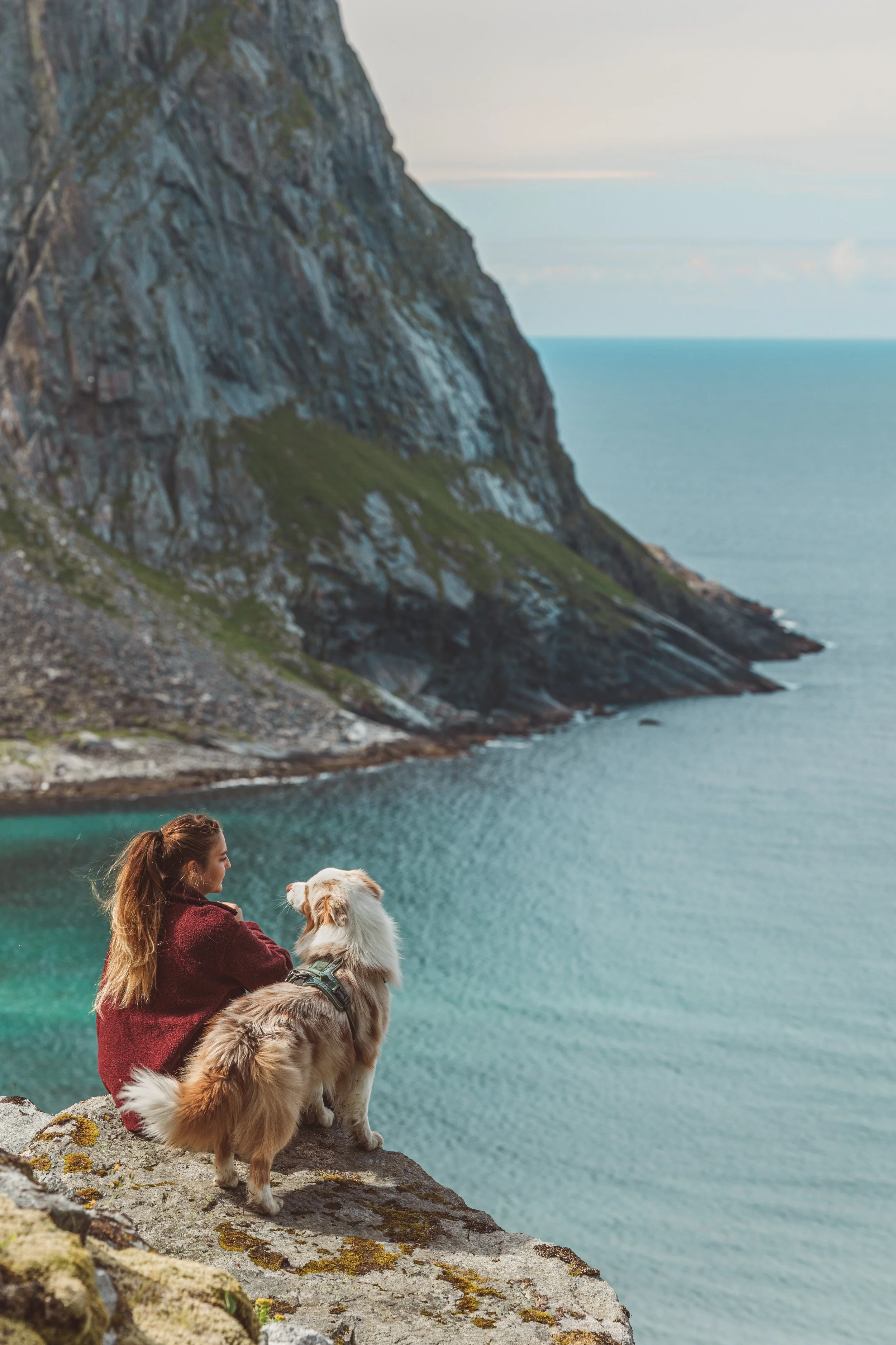 A woman sitting on a rocky cliff with her dog overlooking a body of water and mountainous landscape