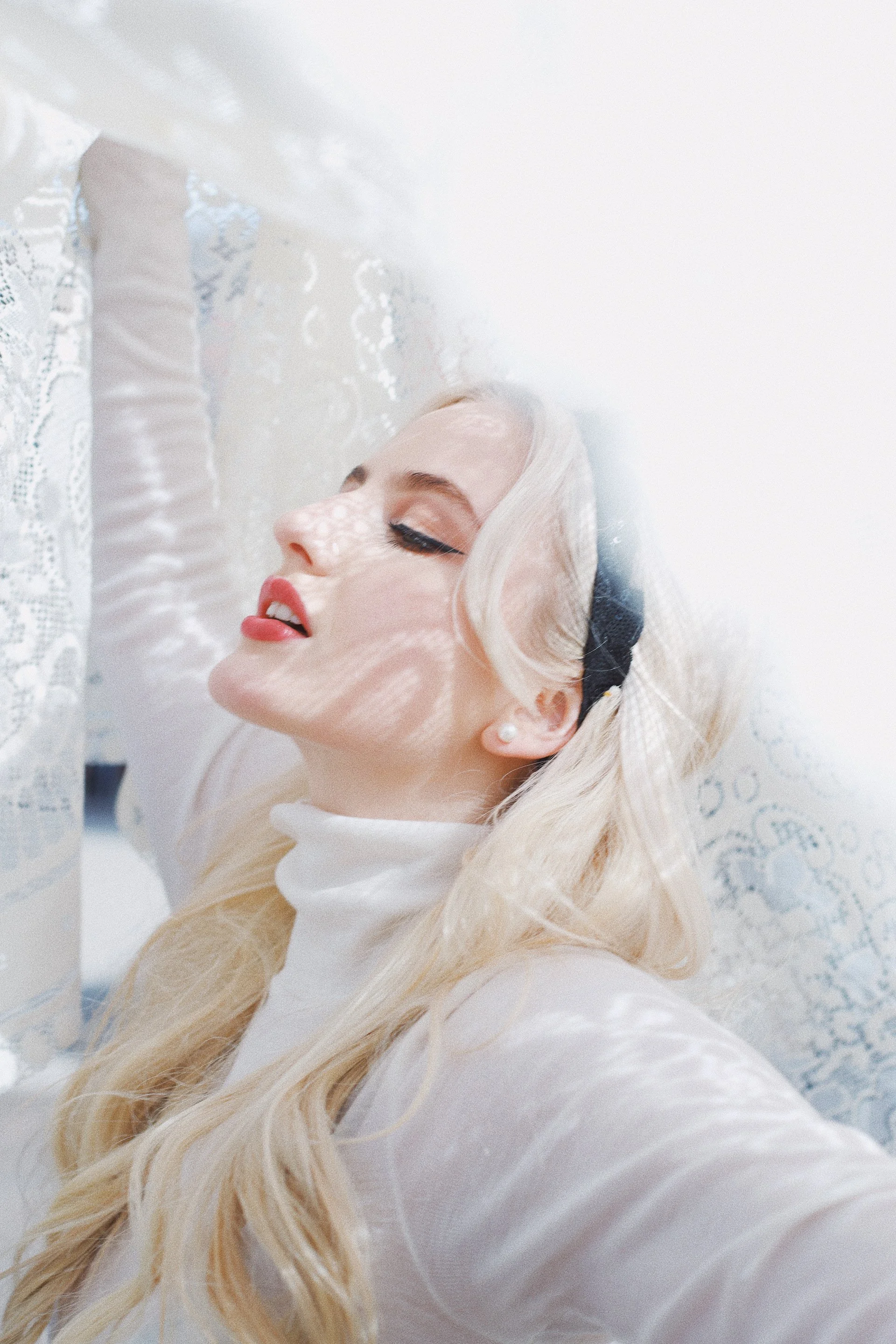 Fanny Avonne with long blonde hair relaxed with eyes closed, leaning against a lace curtain with sunlight creating patterns on her face.