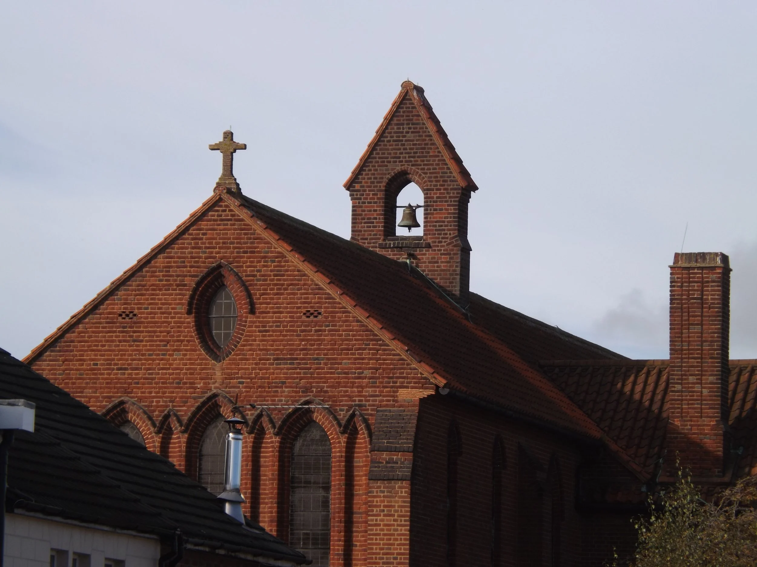 Red brick church with arched windows, a small bell tower with a bell, and a cross on top of the roof, under a partly cloudy sky.