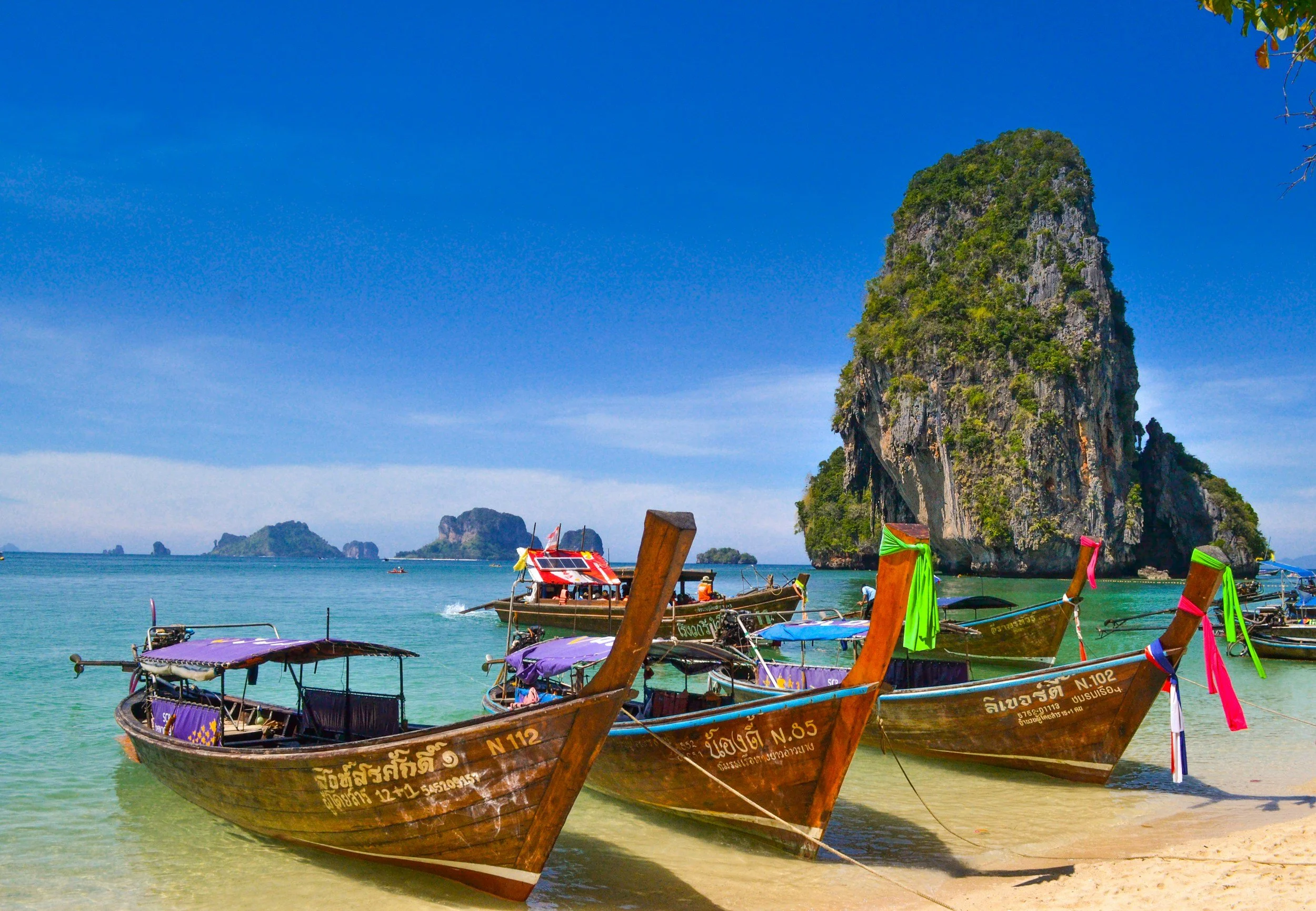 Several traditional wooden longtail boats anchored on a beach with a large limestone island in the background under a clear blue sky.