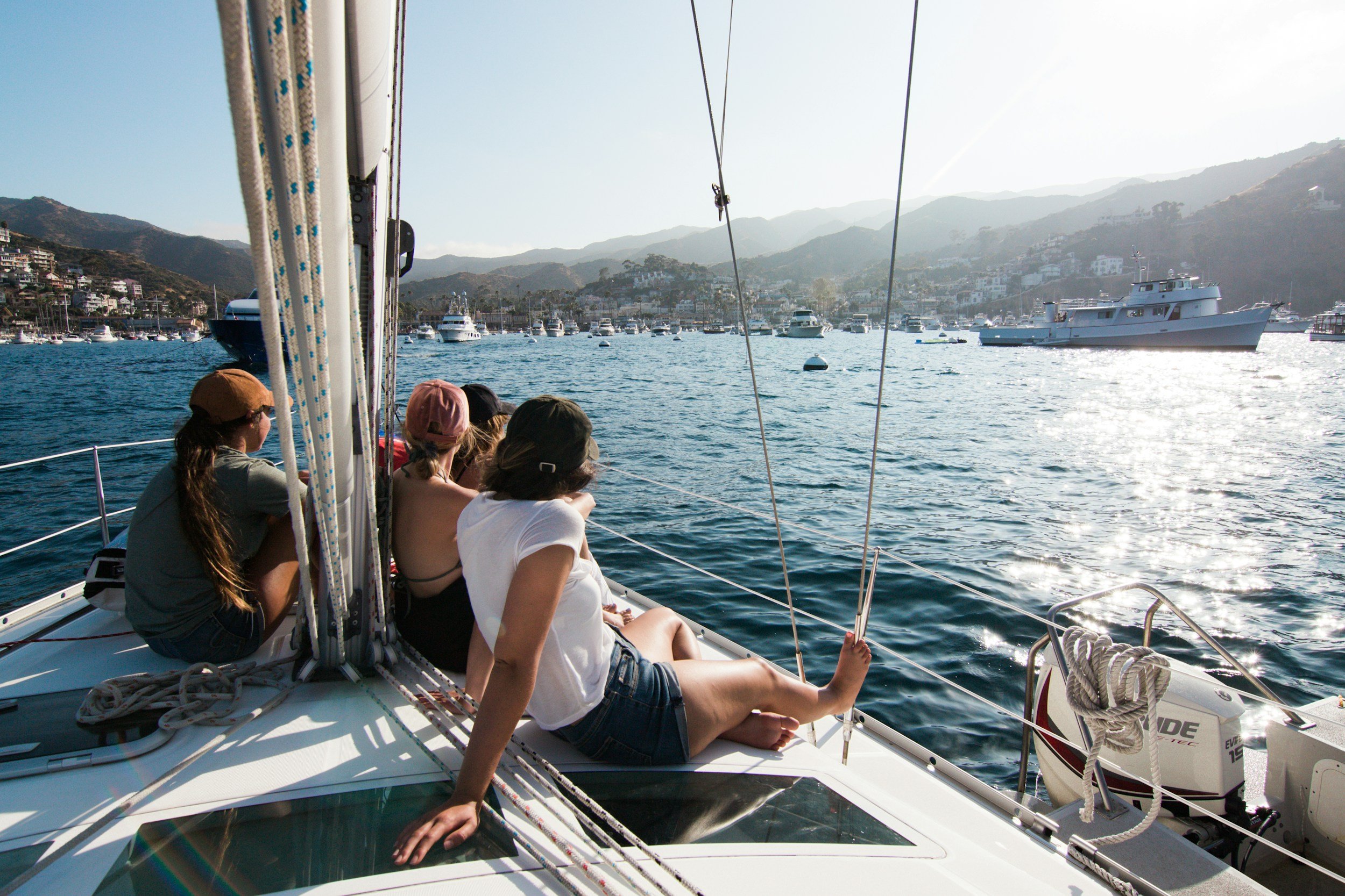A group of people sitting on the deck of a sailboat, looking at the water and distant mountains with boats anchored in a marina, during sunset or sunrise.