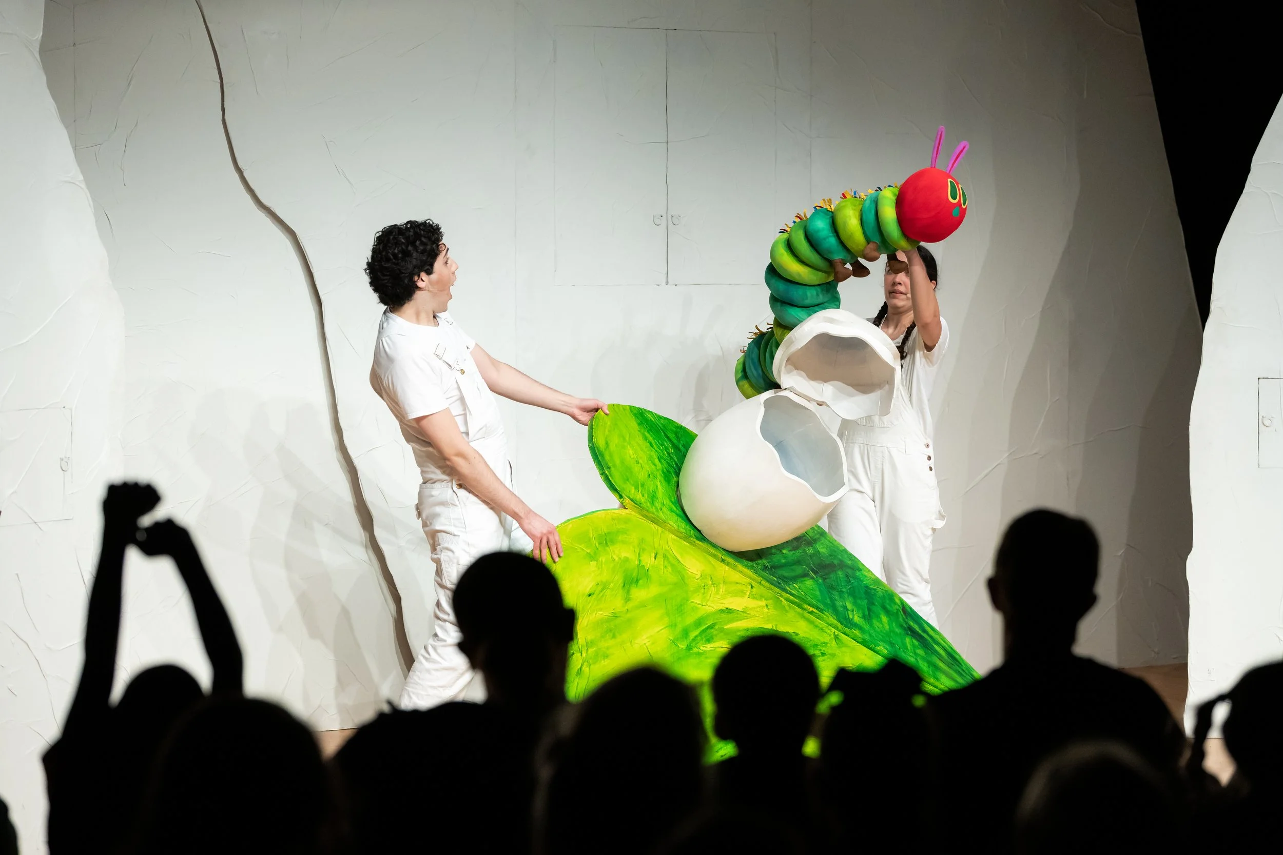 Two performers on stage with a whimsical, colorful caterpillar puppet and a large white eggshell prop, in front of an audience.