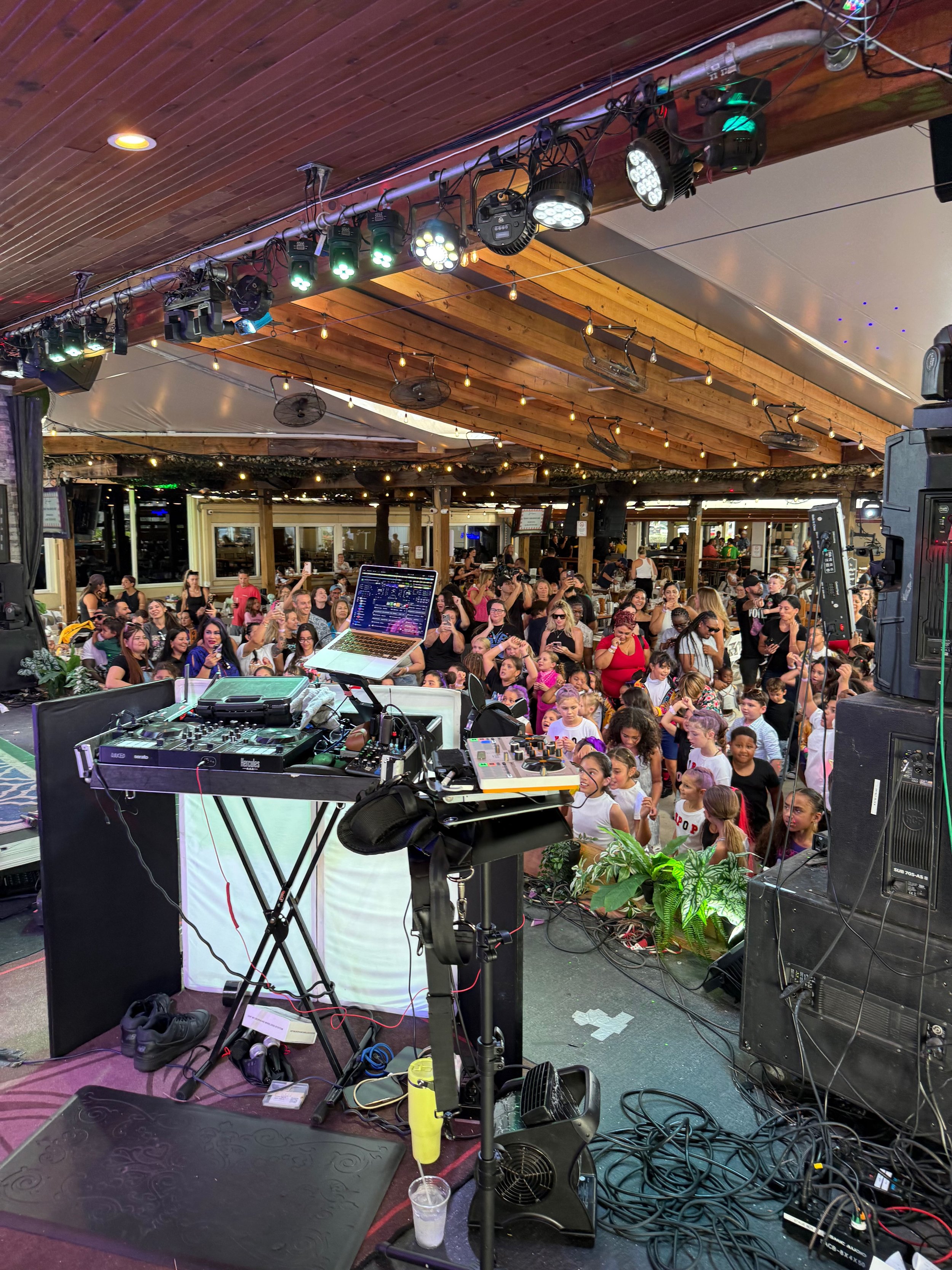 DJ equipment setup in front of a dance floor with a large crowd of people, some dancing, at a music venue with wooden ceiling and string lights.