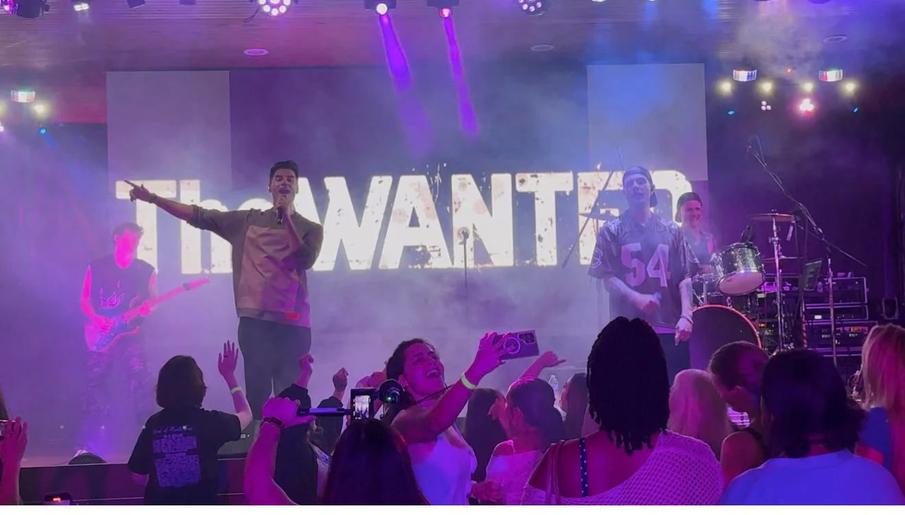 A live concert with a band performing on stage, audience members taking pictures, and a large screen displaying the word 'WANTED' in the background.